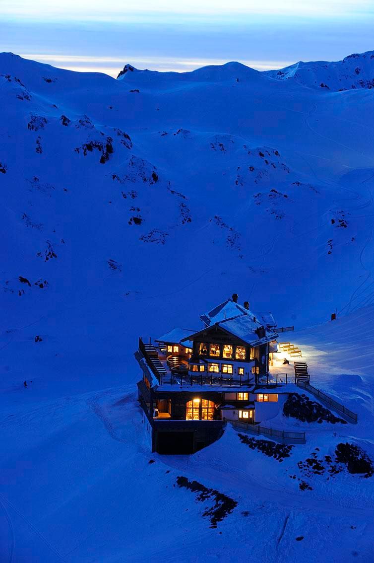 A cozy mountain house in a snowy winter landscape. The windows radiate warm light into the cold, blue surroundings.