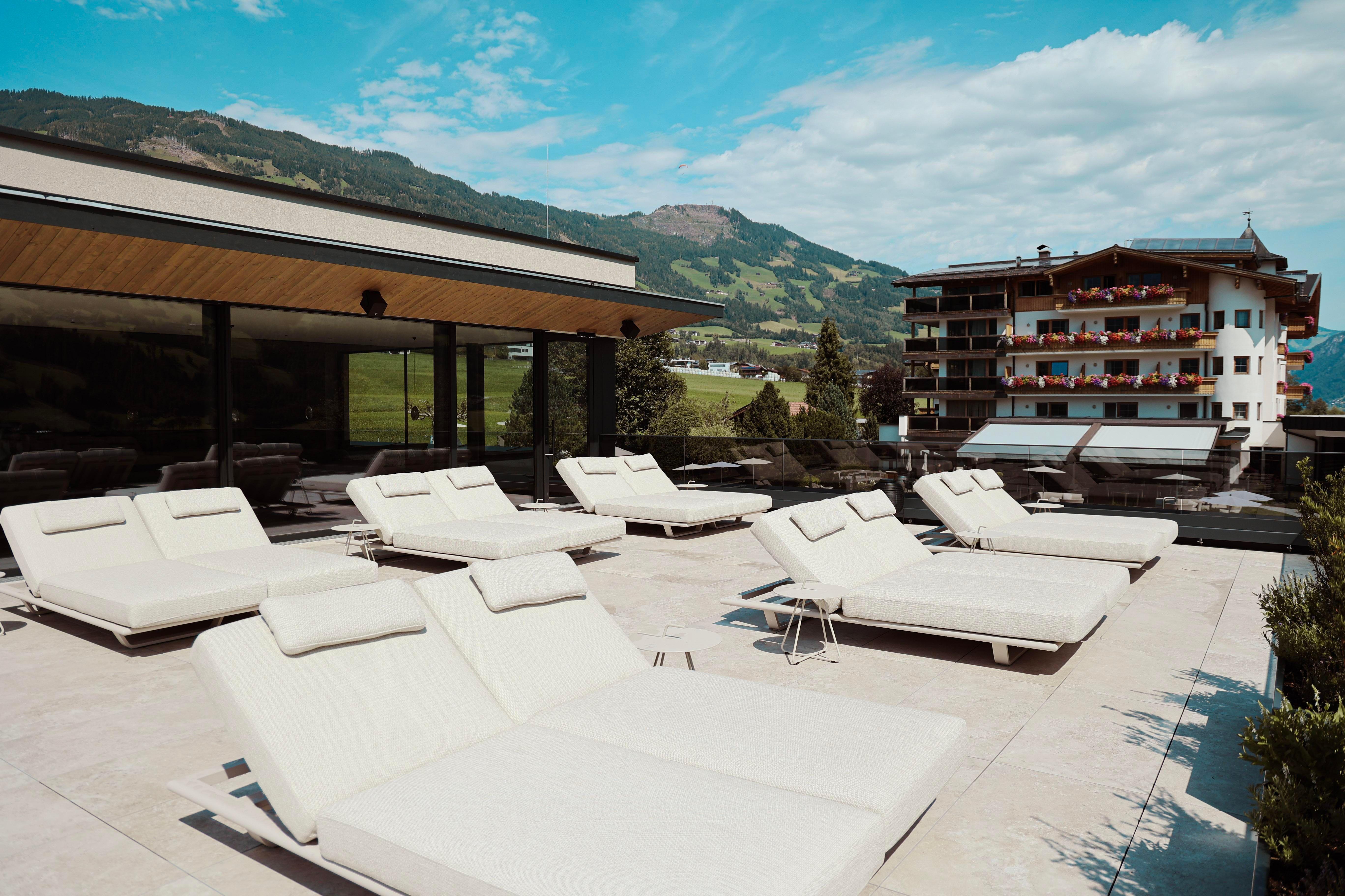A modern rooftop terrace with white loungers and a view of the surrounding mountains. The sky is blue with a few clouds.