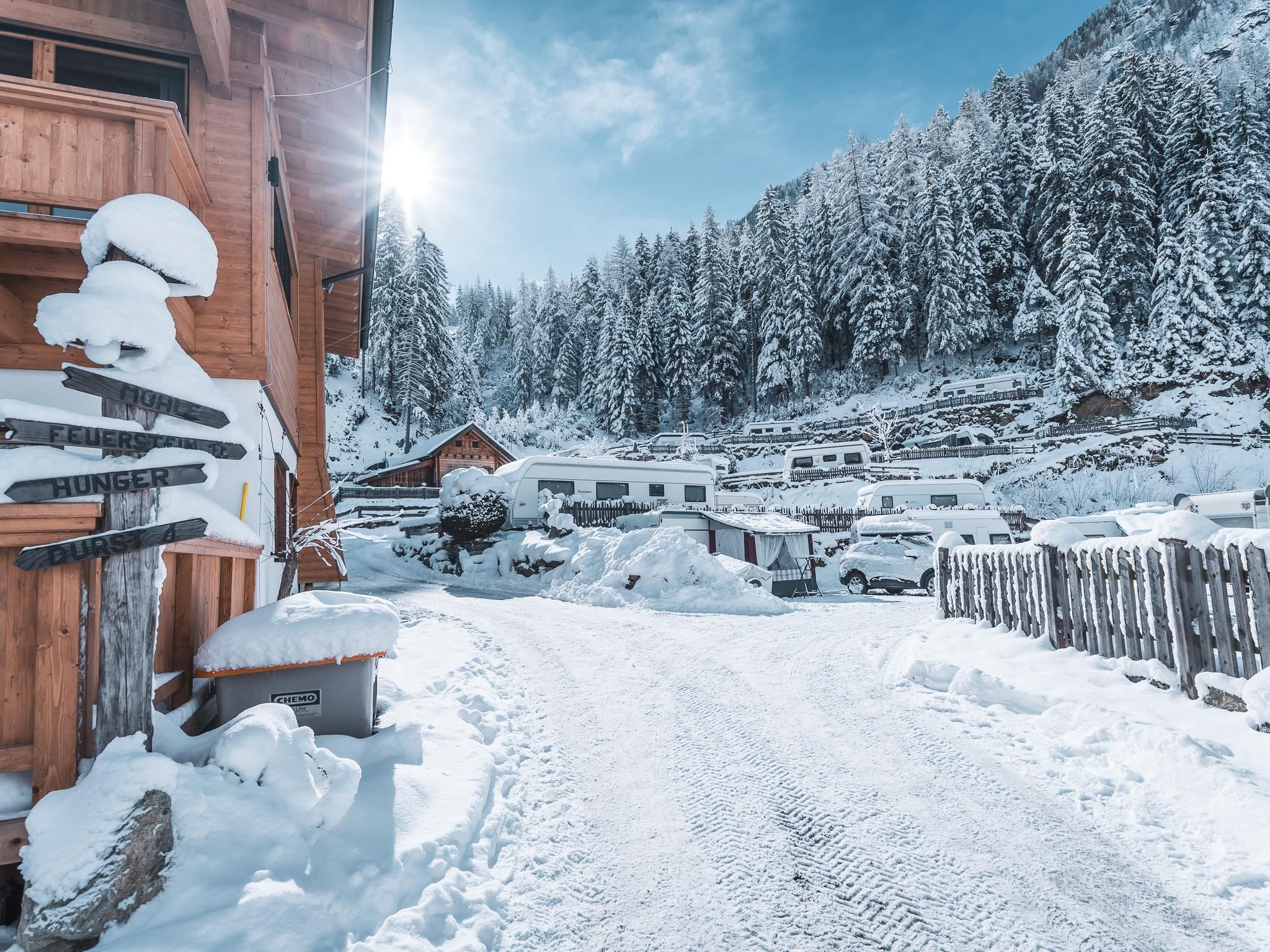 A snow-covered landscape with tall fir trees and a clear sky. In the foreground, a snowy road and wooden buildings can be seen.