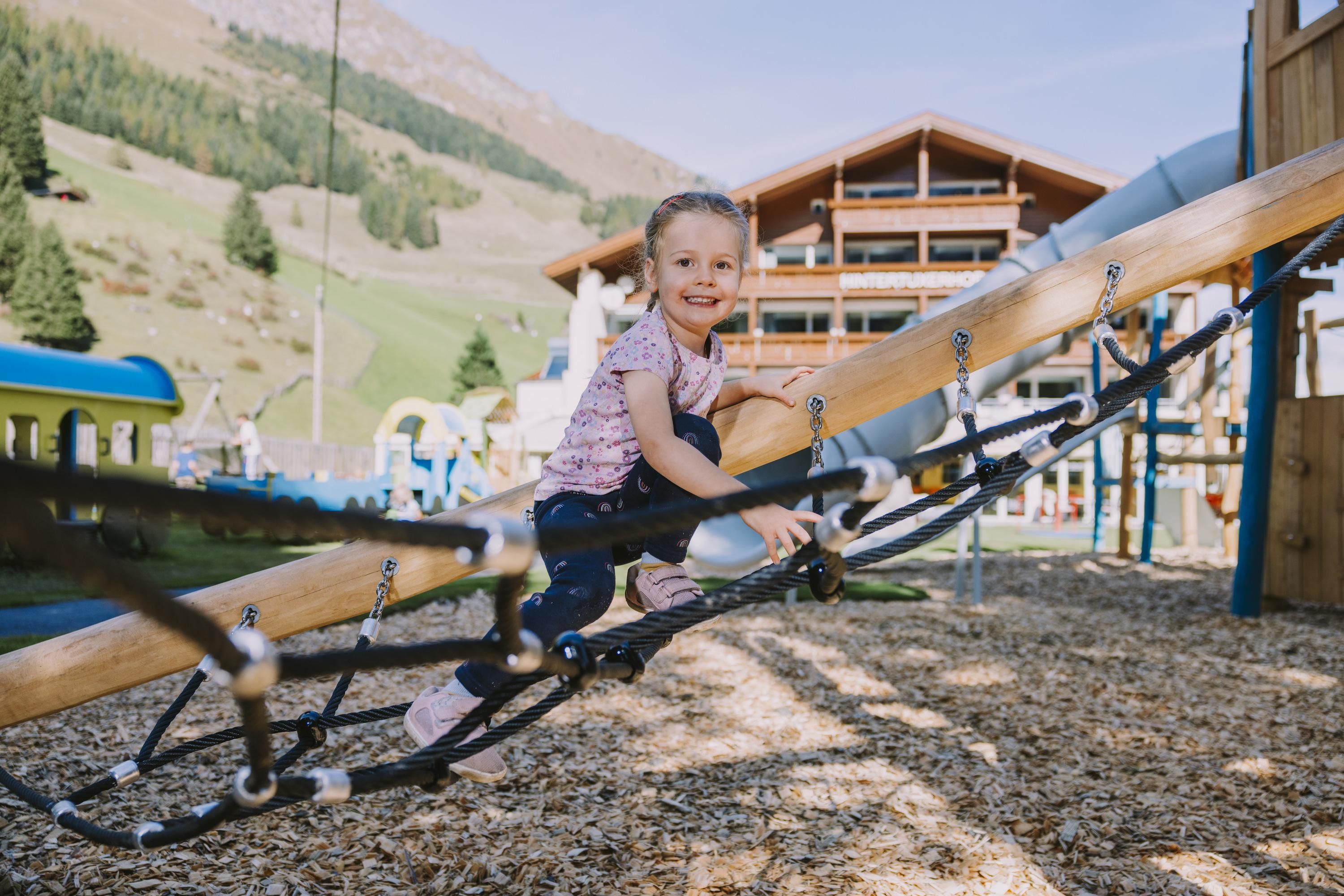 A happy child is playing on a playground. In the background, green hills and a building can be seen.