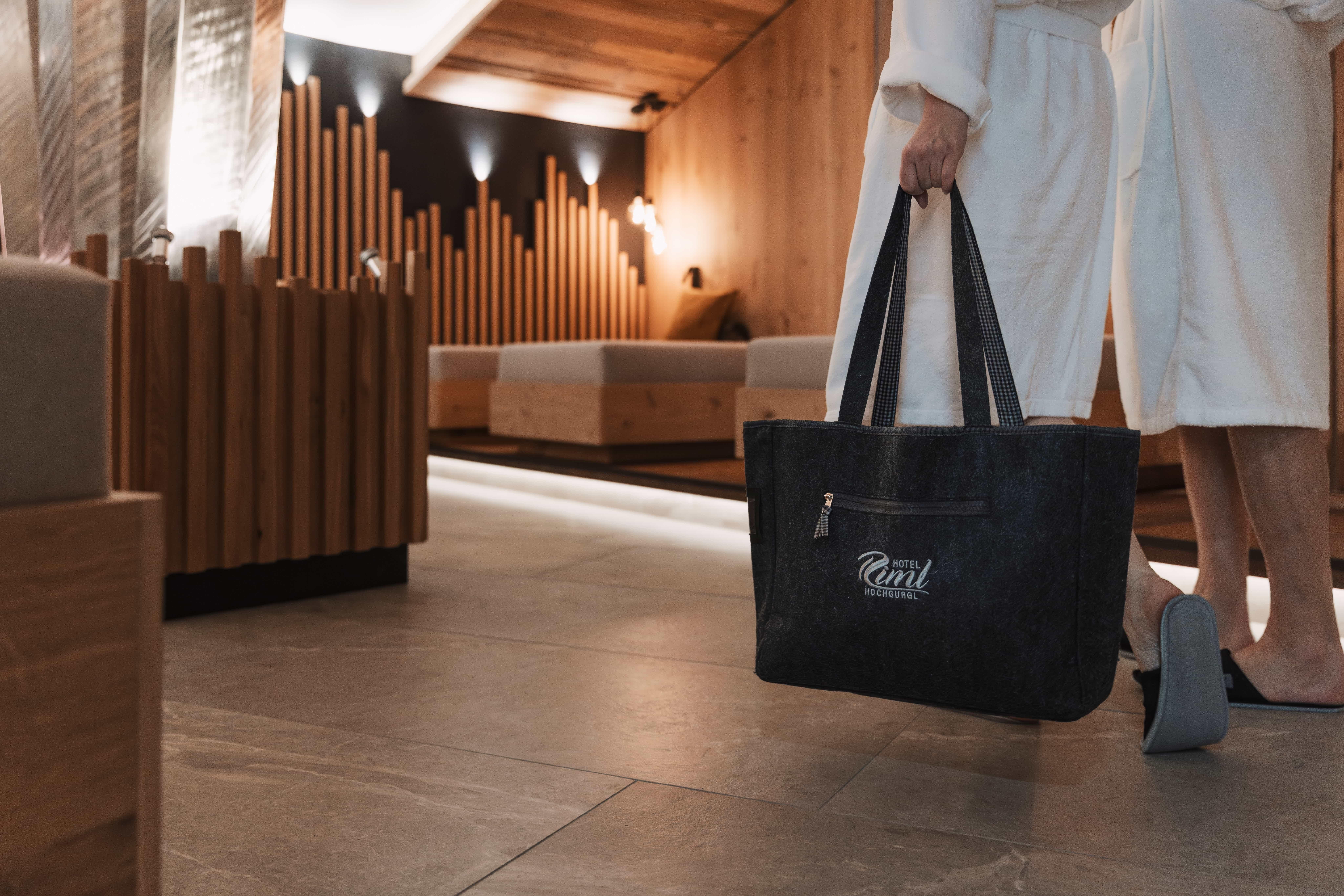 A person in a bathrobe is carrying a large bag in a modern, elegant spa area. The room is cozily designed with wood and soft lighting.