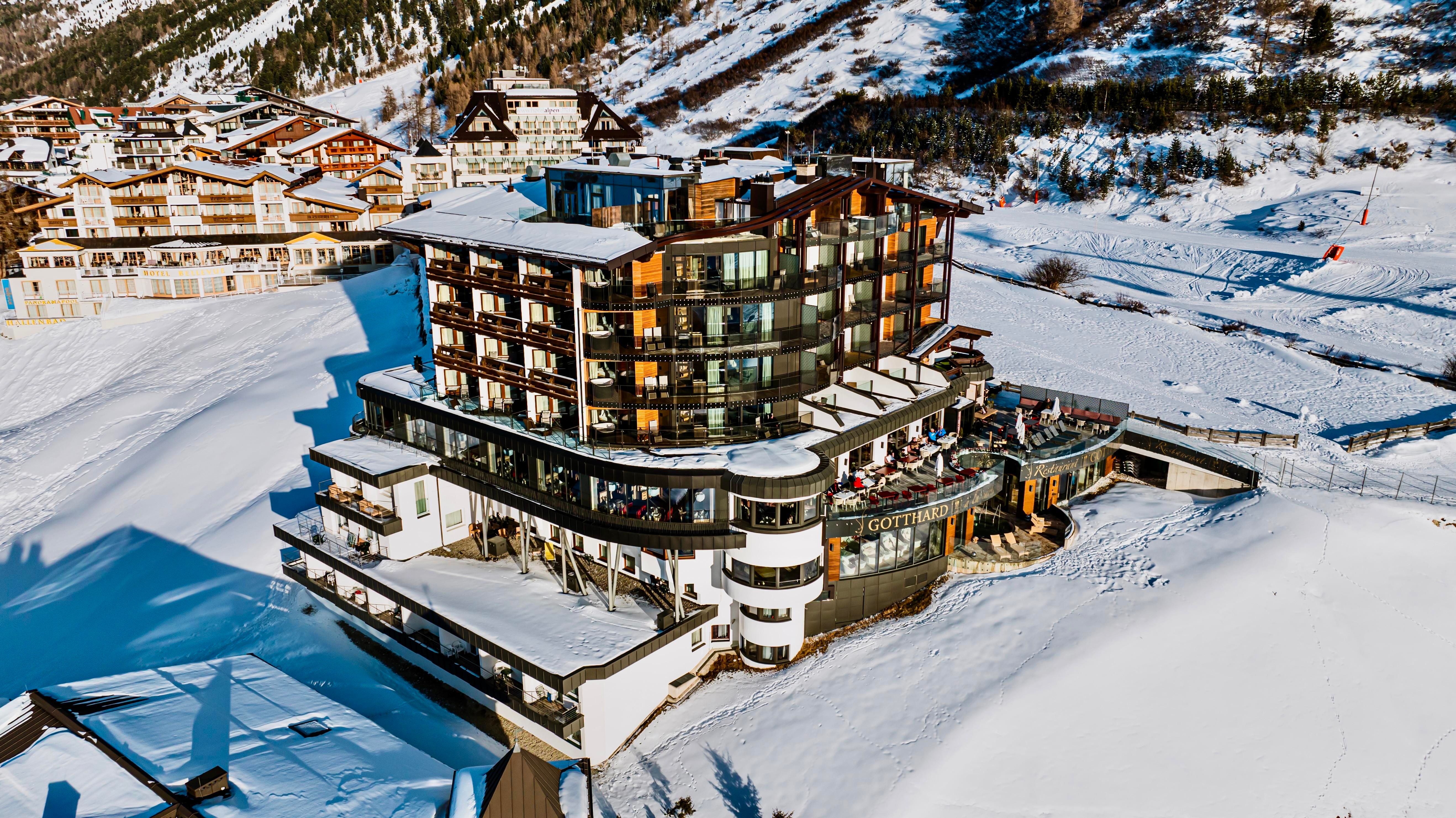 A modern hotel in the snow with large windows and a cozy restaurant. In the background, snow-covered mountains and other buildings extend.