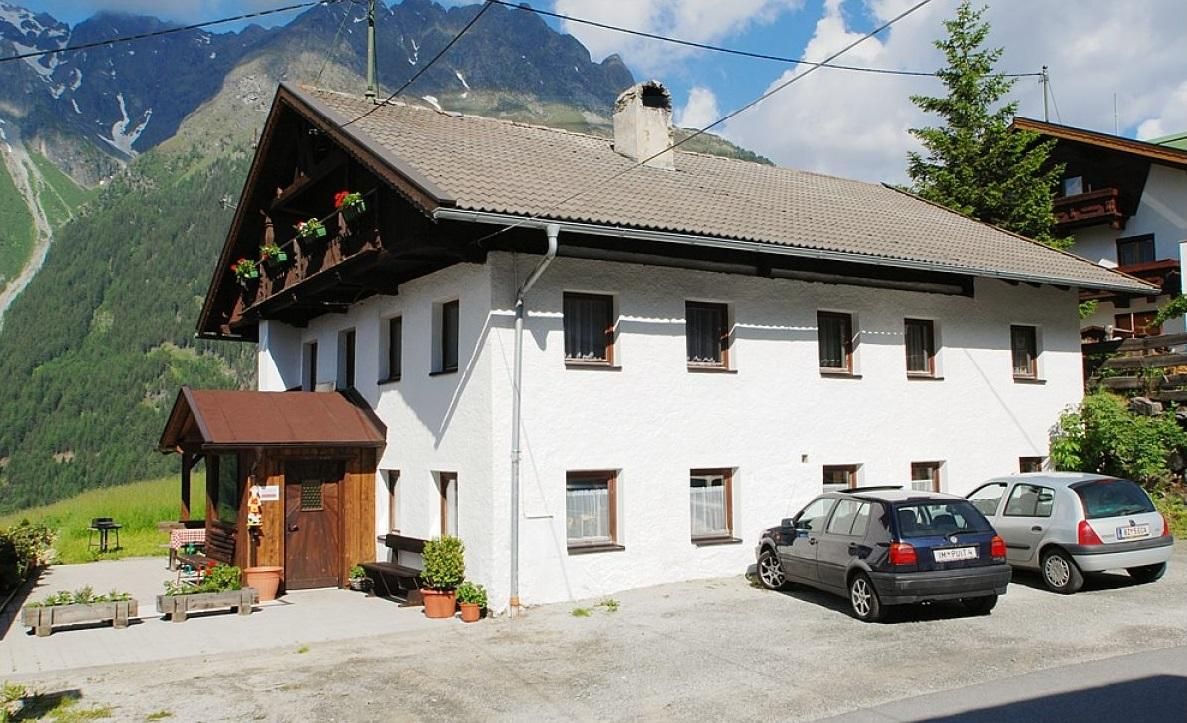 A cozy, two-story house in alpine style, surrounded by mountains. Several cars are parked in front of the house, and there are flower boxes on the windows.
