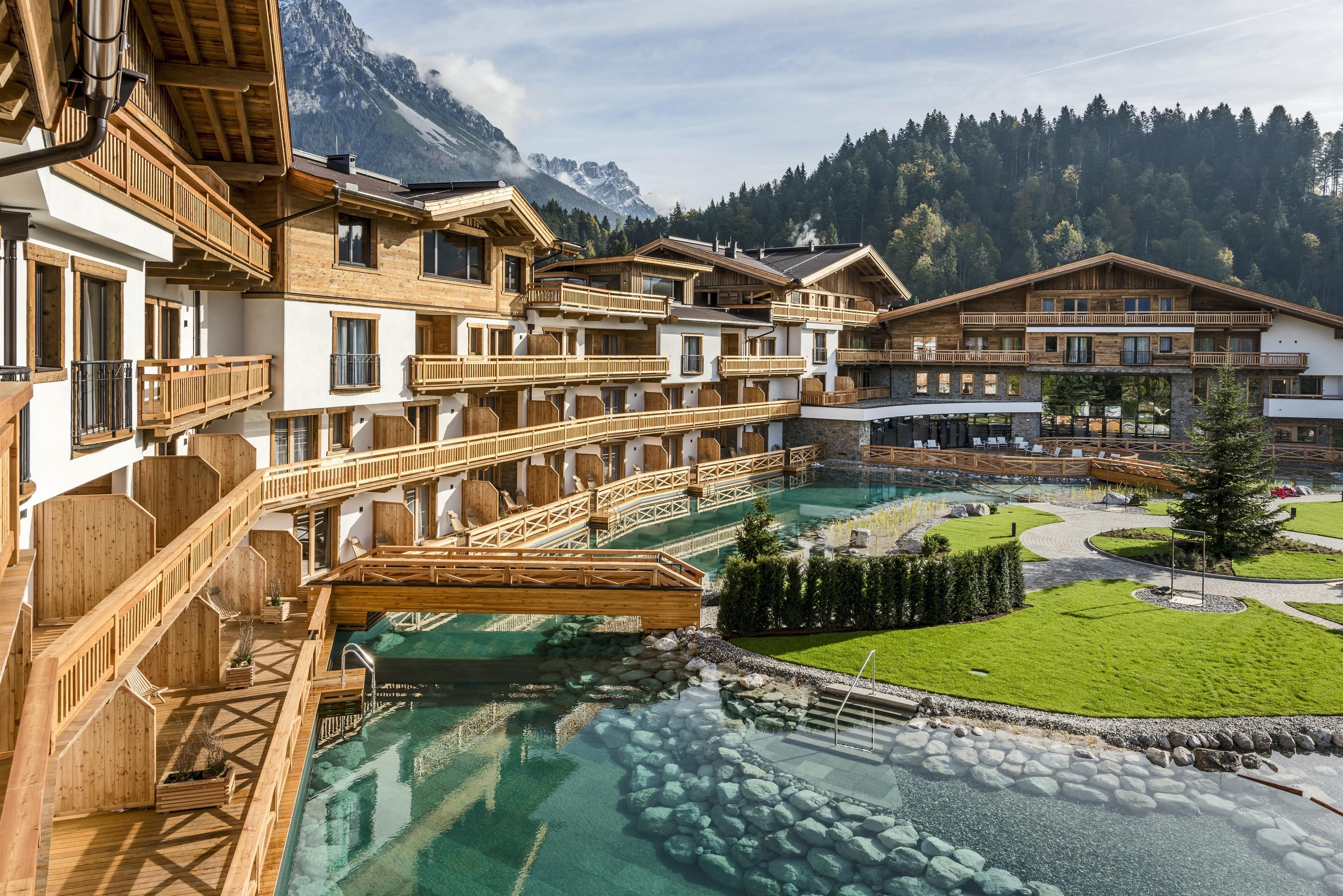A beautiful wellness resort with wooden architecture and a clear water stream. In the background, the mountains and a green area with grass and plants can be seen.