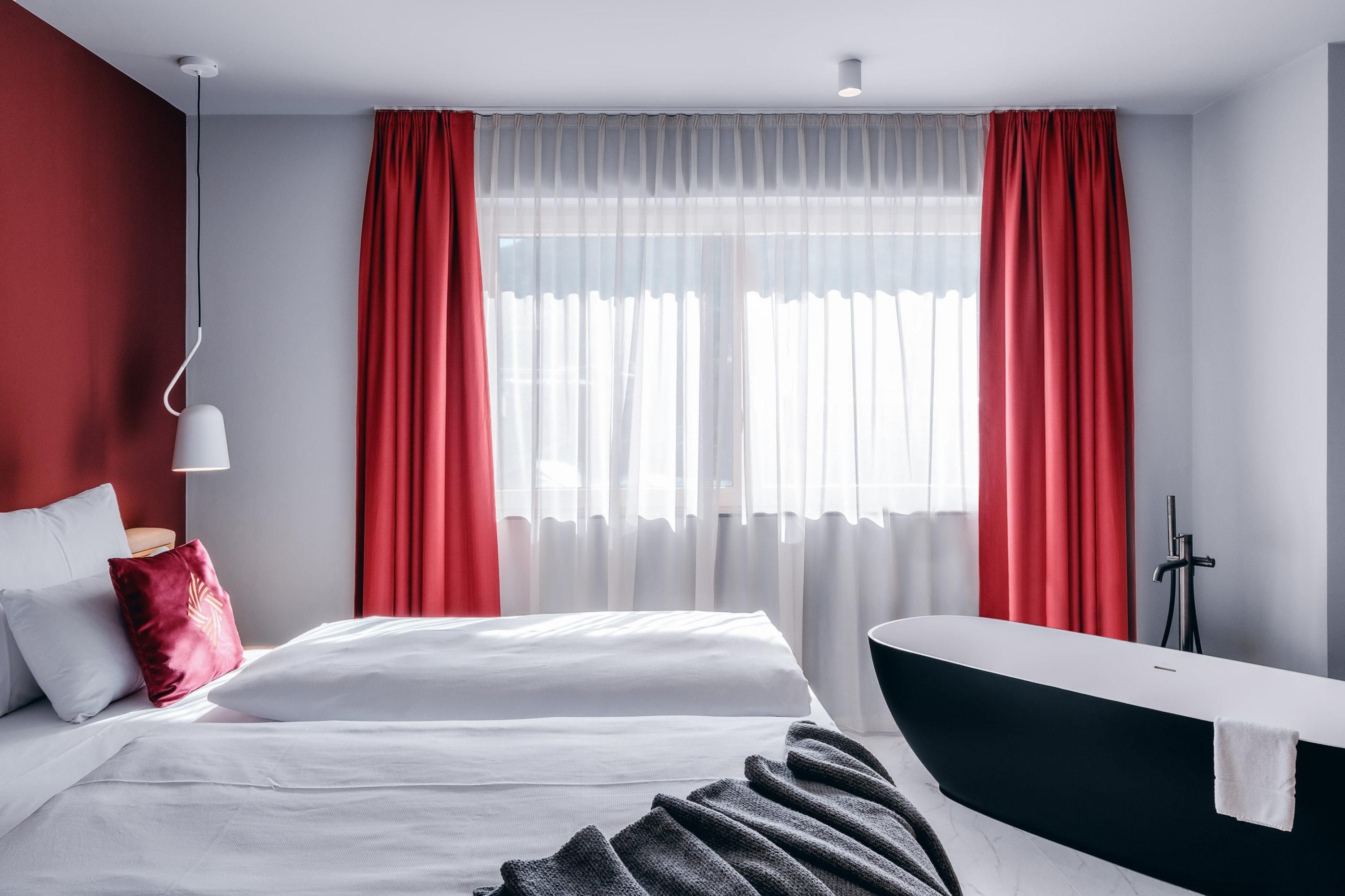 A modern hotel room with a large window and red curtain. The room features a comfortable bed and a freestanding bathtub.