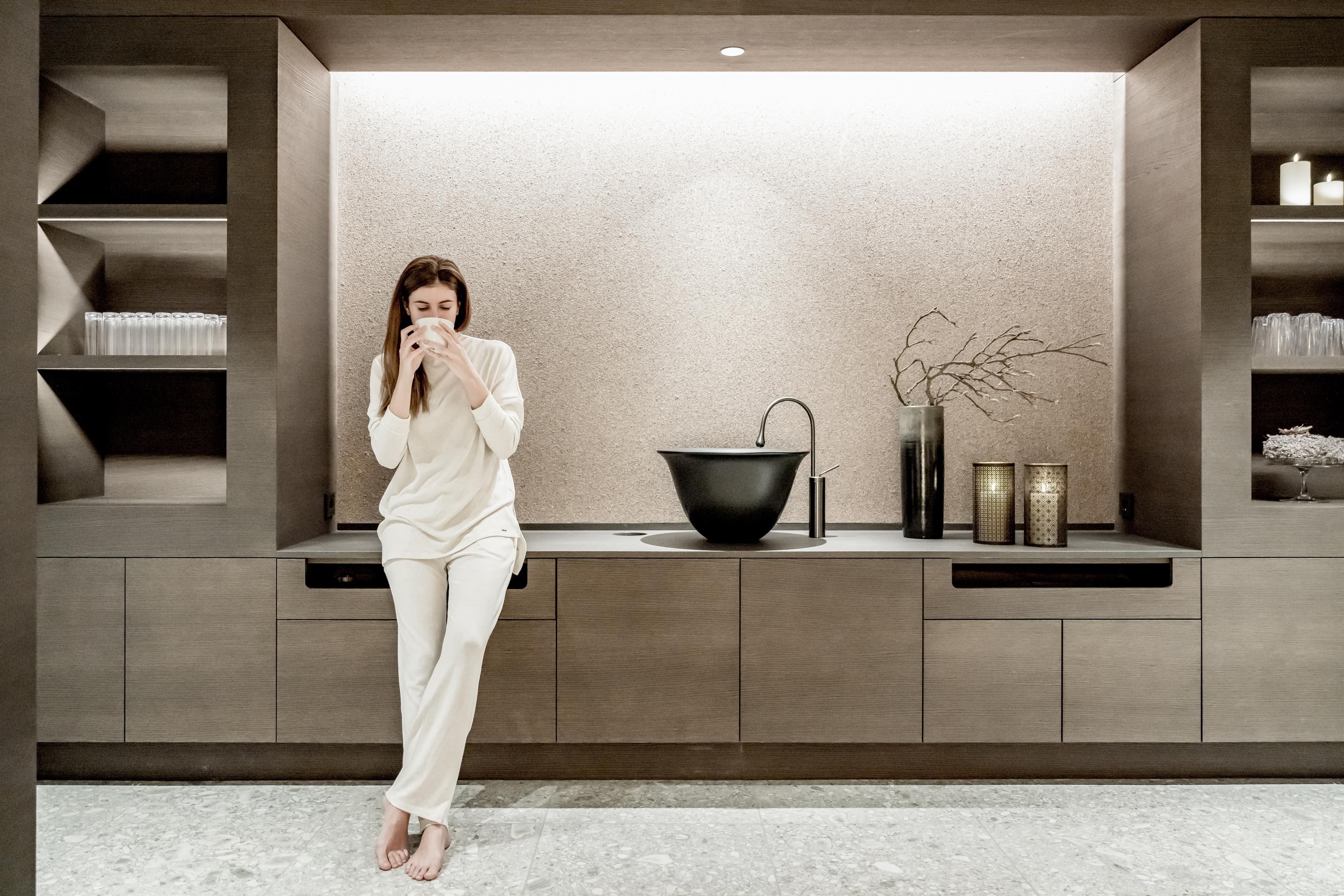 A woman in comfortable clothing stands at a modern, stylish kitchen countertop. In the background, an elegant sink and decorative elements are visible.