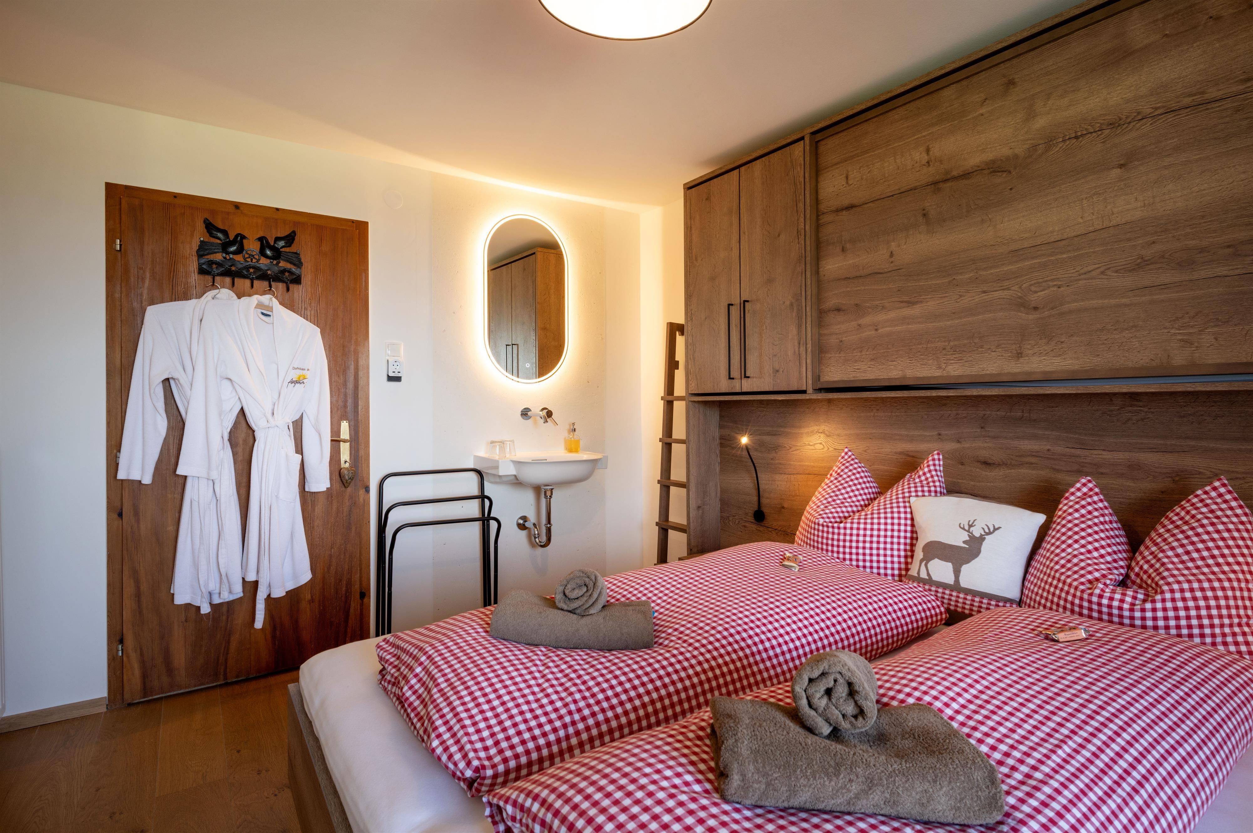 A cozy bedroom with two single beds and a red-white checked pattern. There is a mirror, a small wash area, and bathrobes hanging on the door.