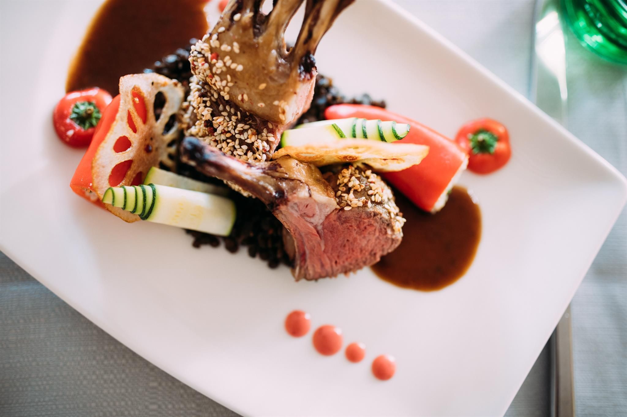 An artfully arranged dish featuring lamb, accompanied by colorful vegetables and a spicy sauce. The whole is presented on a white plate and sprinkled with sesame.