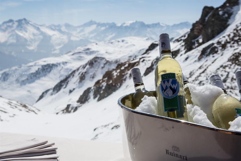 An elegant ice bath with bottles of wine stands in the snow. In the background, majestic mountain peaks stretch under a clear sky.
