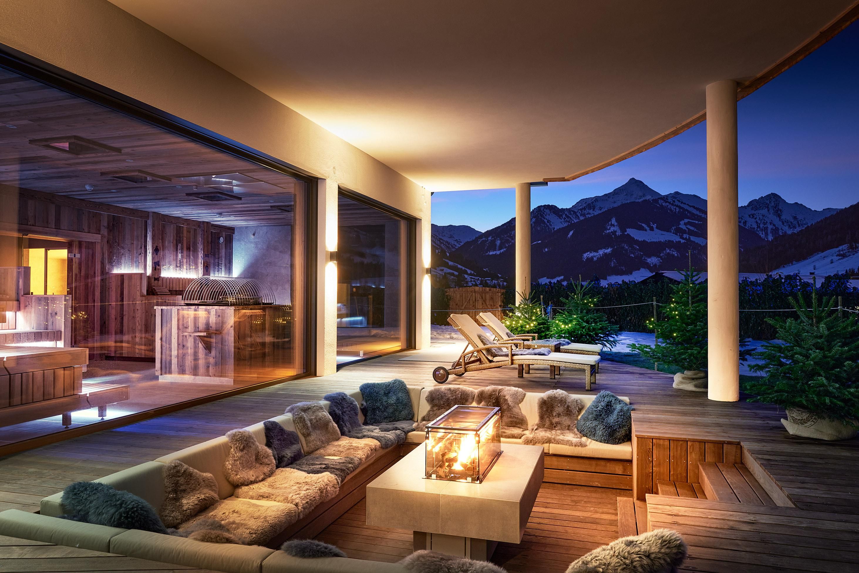 A stylish terrace with comfortable seating and a fireplace. In the background, mountains are visible under the evening sky.