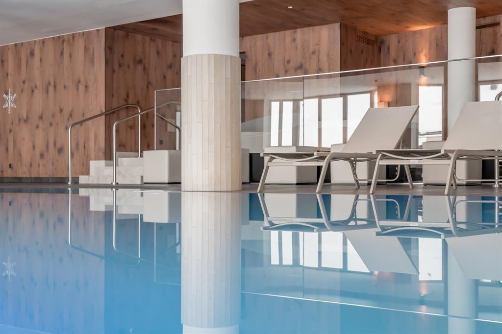 An elegant indoor swimming pool with clear blue water and modern loungers. The walls are in warm wood and neutral tones.