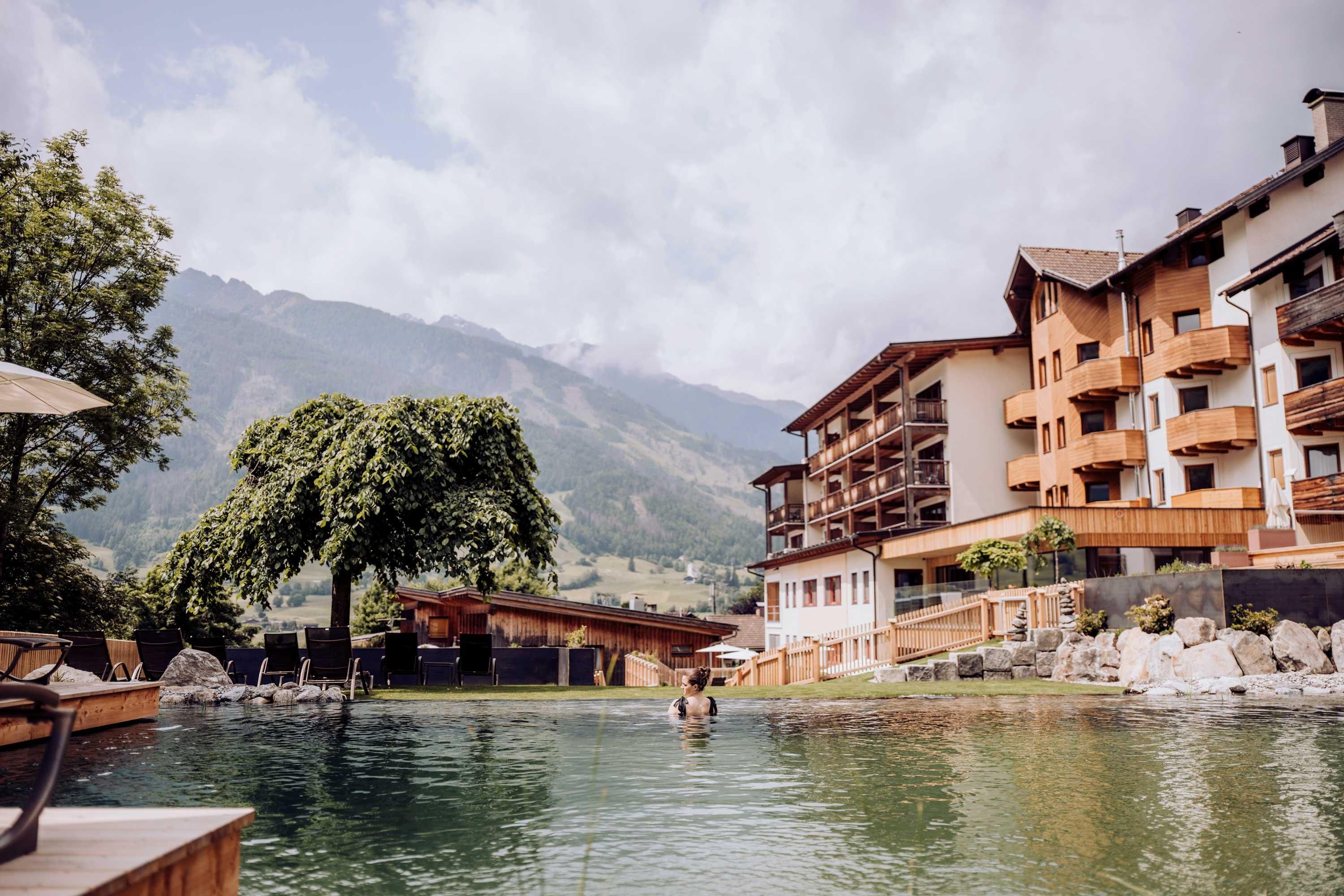 The garden of the Naturhotel Outside in Matrei in Osts Tirol by the water with mountains in the background. In the foreground, a person is swimming in the clear water, surrounded by nature.