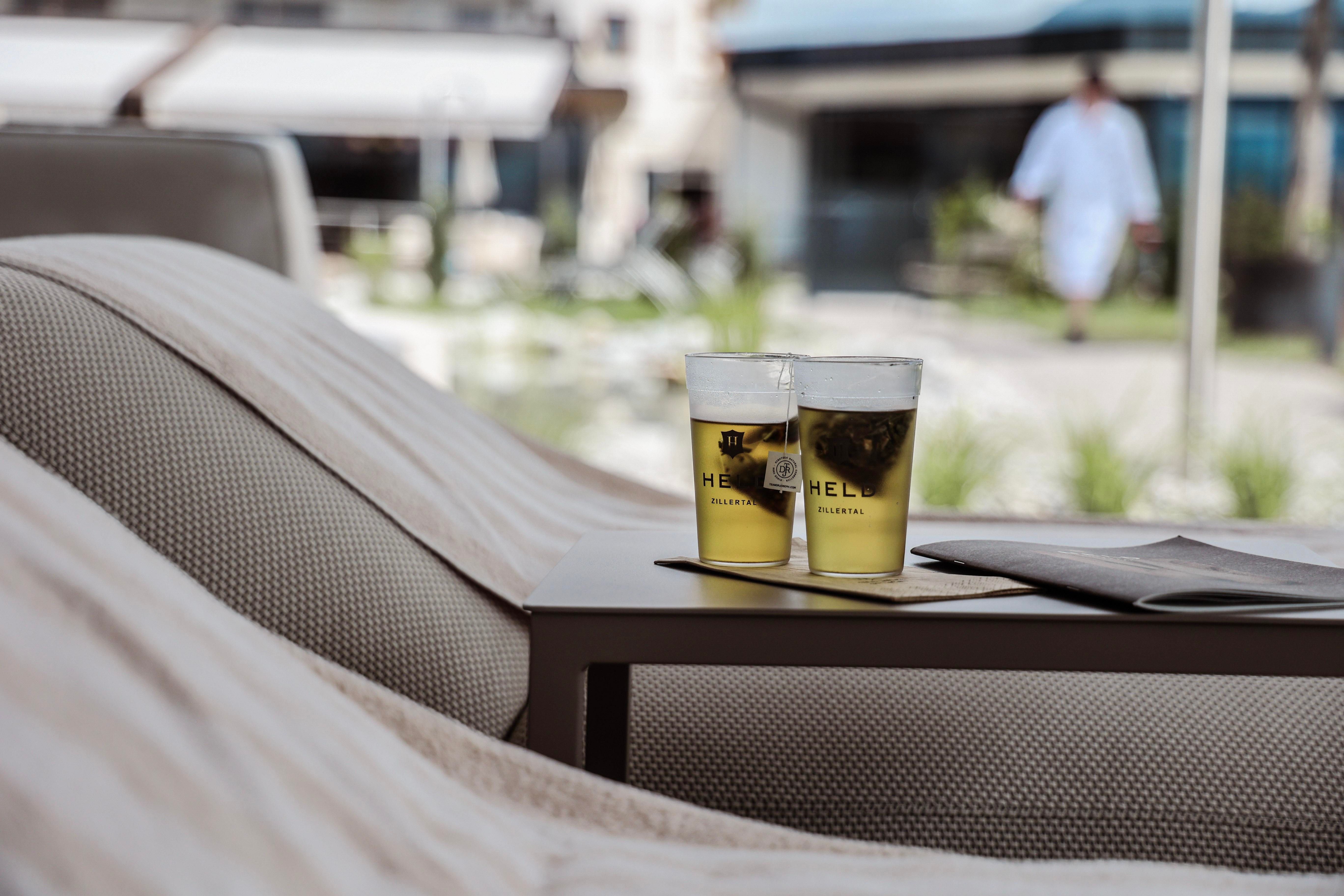 Two glasses with drinks are on a table, surrounded by a relaxing atmosphere. In the background, there is a calm environment with plants and a person in a bathrobe visible.