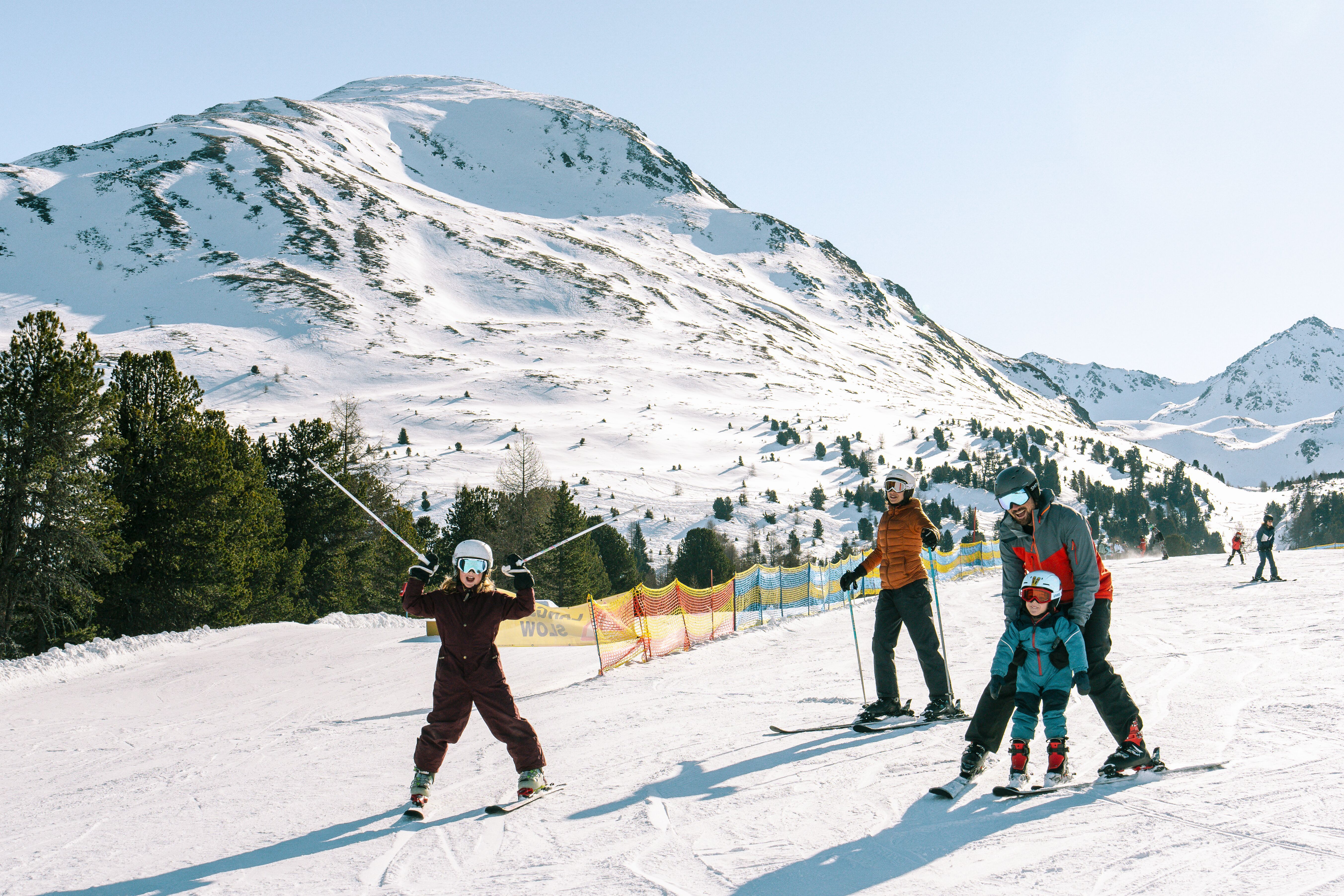 Man sieht eine Familie auf der Skipiste in Nauders. Der Vater lernt dem jüngeren Kind Skifahren, die Mutter und das ältere Kind haben Spaß auf der blauen Piste.