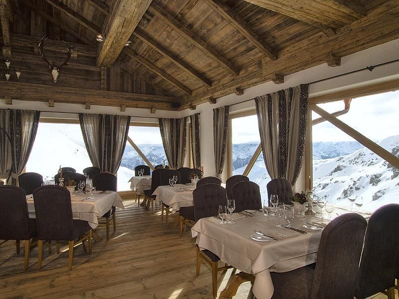 An elegant restaurant with a wood-paneled ceiling and large windows offering a view of snow-capped mountains. The tables are set with white tablecloths and fine dishes.