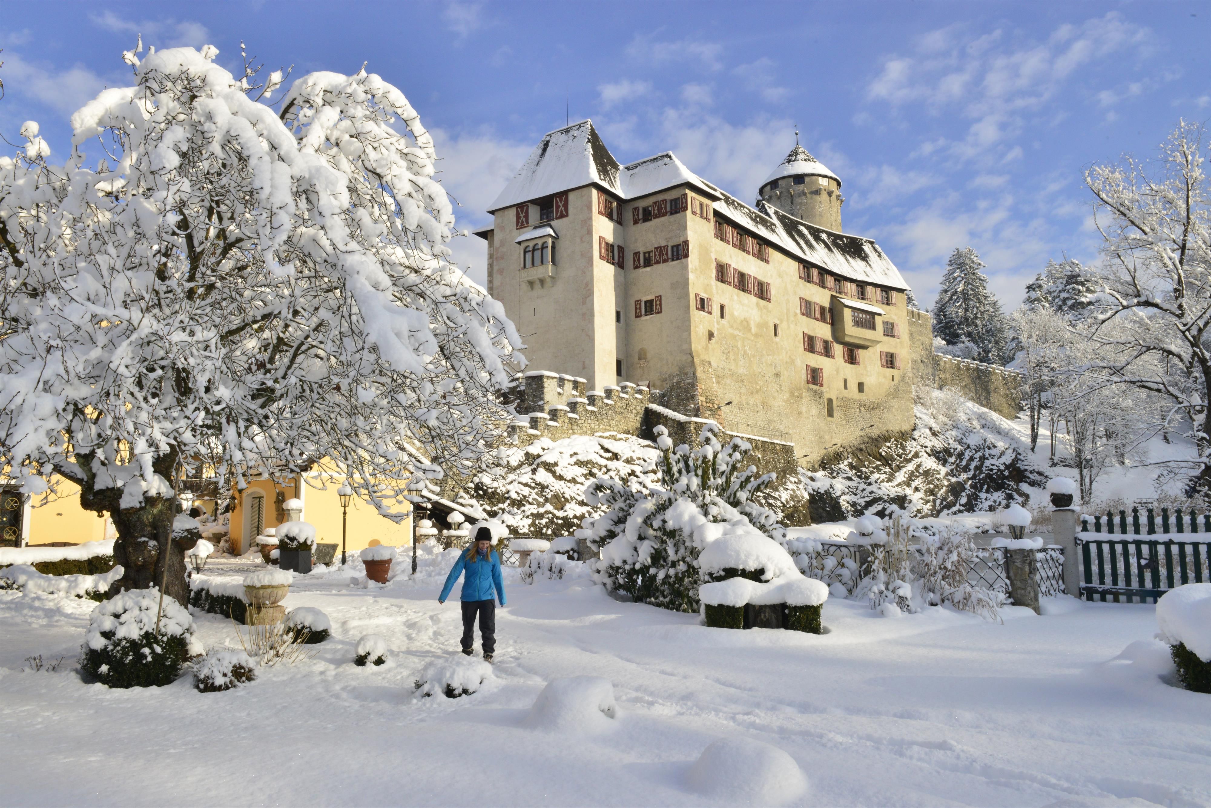 A snow-covered castle surrounded by a wintry landscape. A person is walking through the fresh snow.