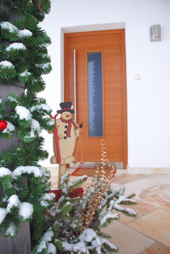 A festively decorated entrance door with a snowman and a green fir tree. The ground is adorned with snow and plants.