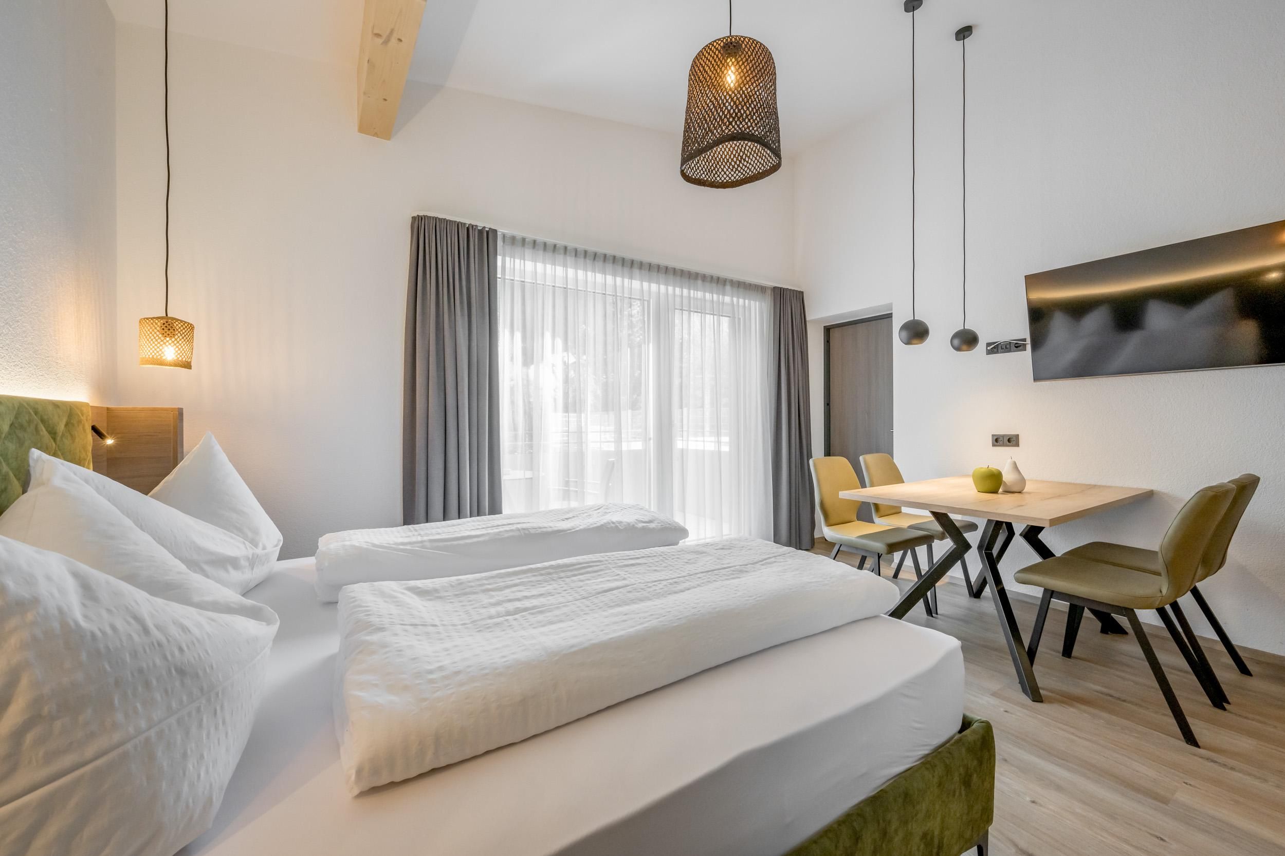 A modern room with two beds and a table. Large windows provide plenty of natural light.