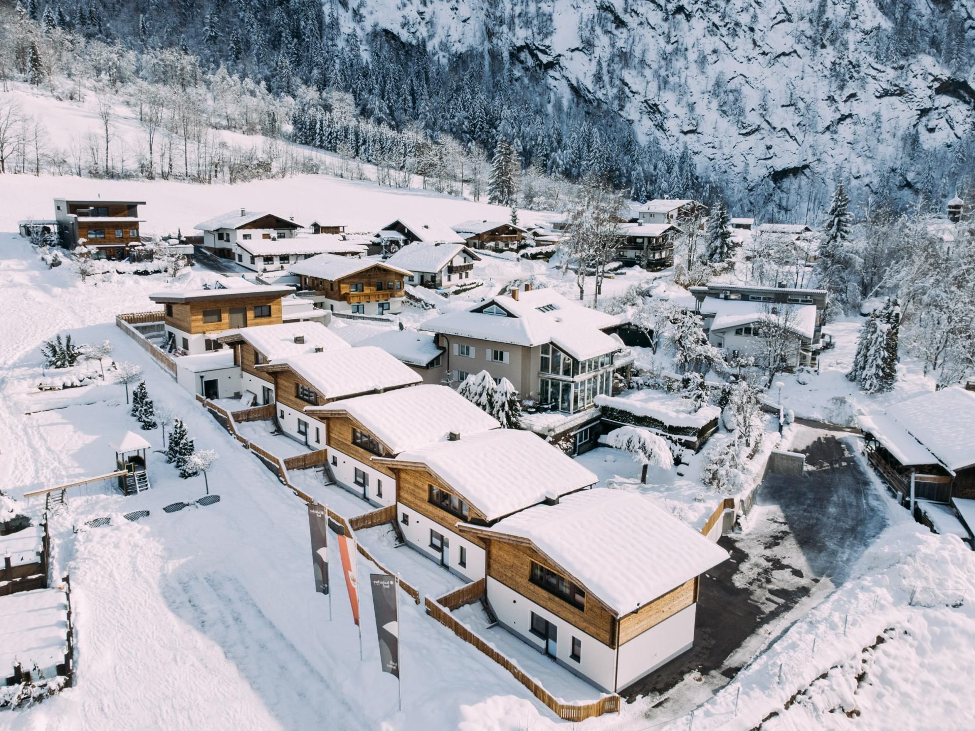 A winter landscape with several chalets covered in snow. In the background, mountains and additional houses can be seen.