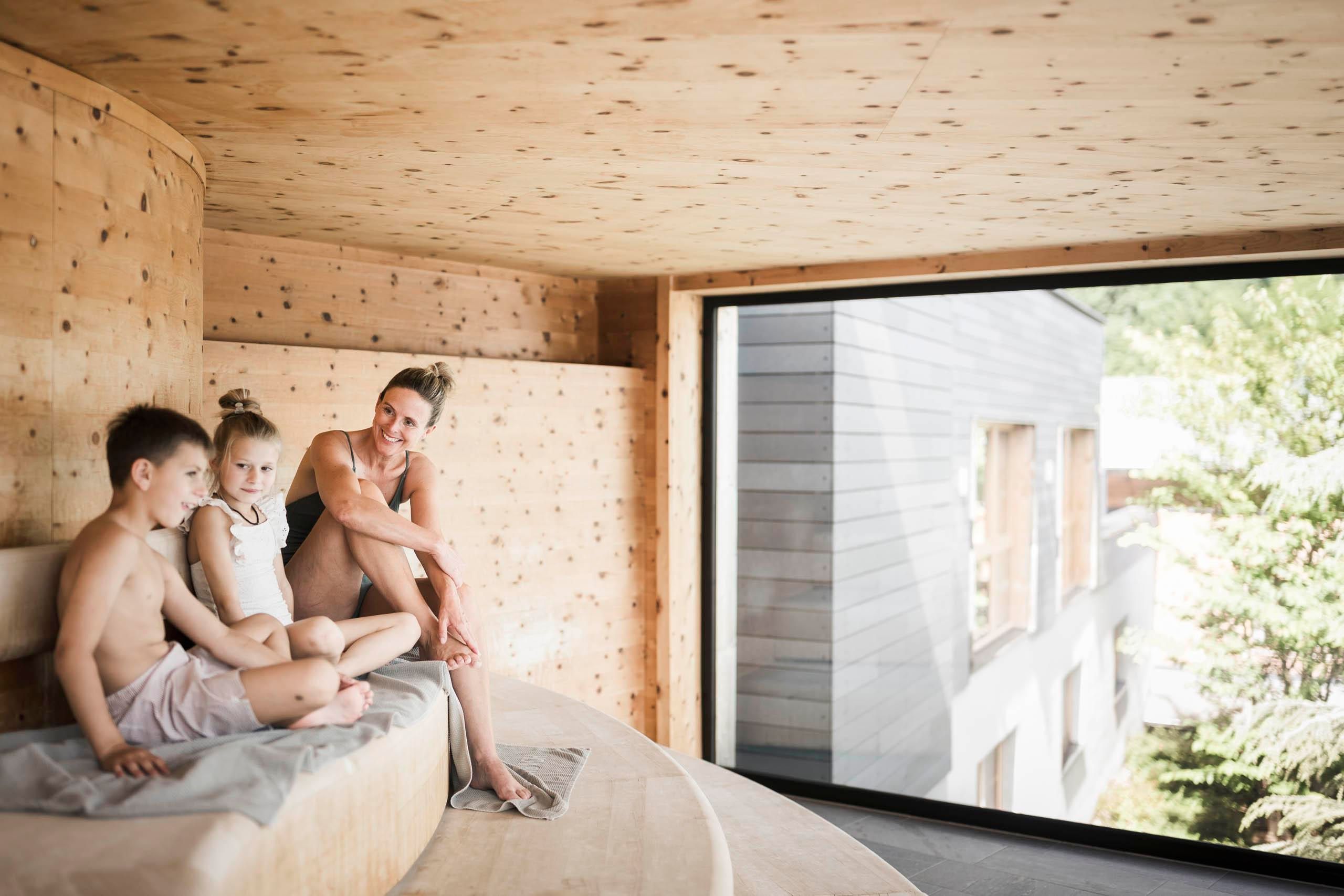 A family is relaxing in a modern, bright room with wood paneling. They are sitting on a comfortable seating area and enjoying the view through large windows.