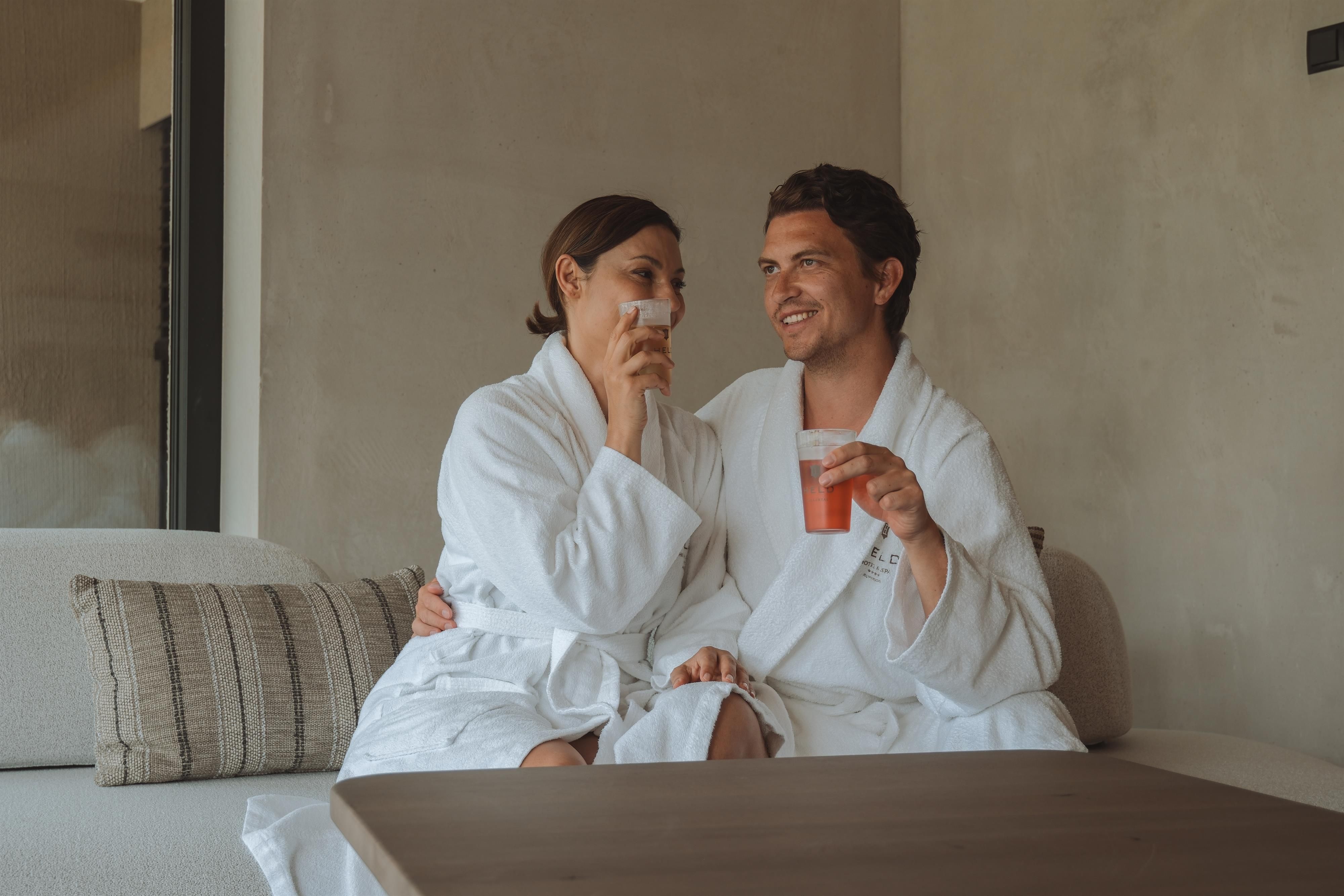 A couple in white bathrobes is sitting comfortably on a couch. They are enjoying drinks and having a relaxed time together.