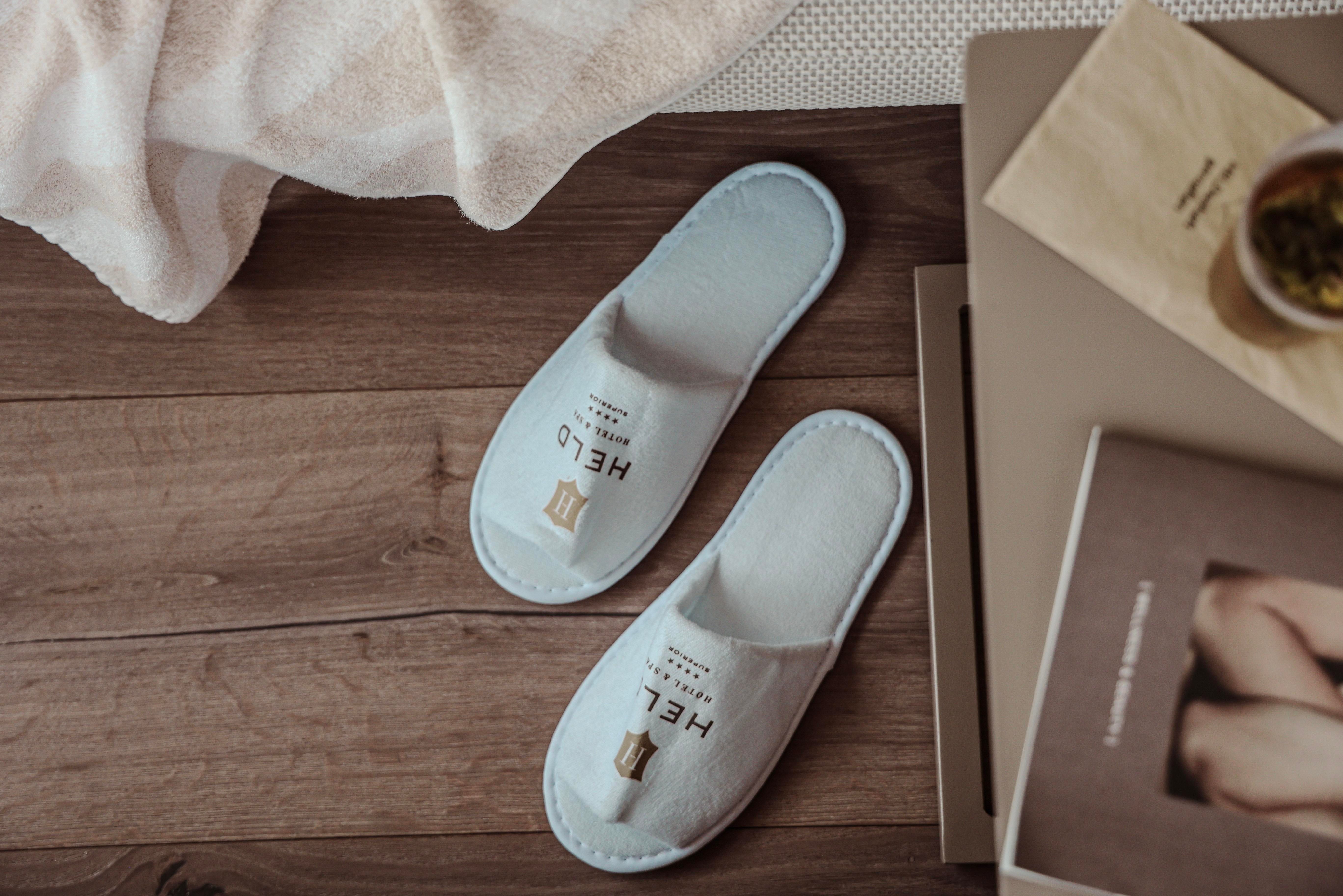 Two slippers with the inscription "HELLO" are lying on a wooden floor. In the background, a towel and some books can be seen.