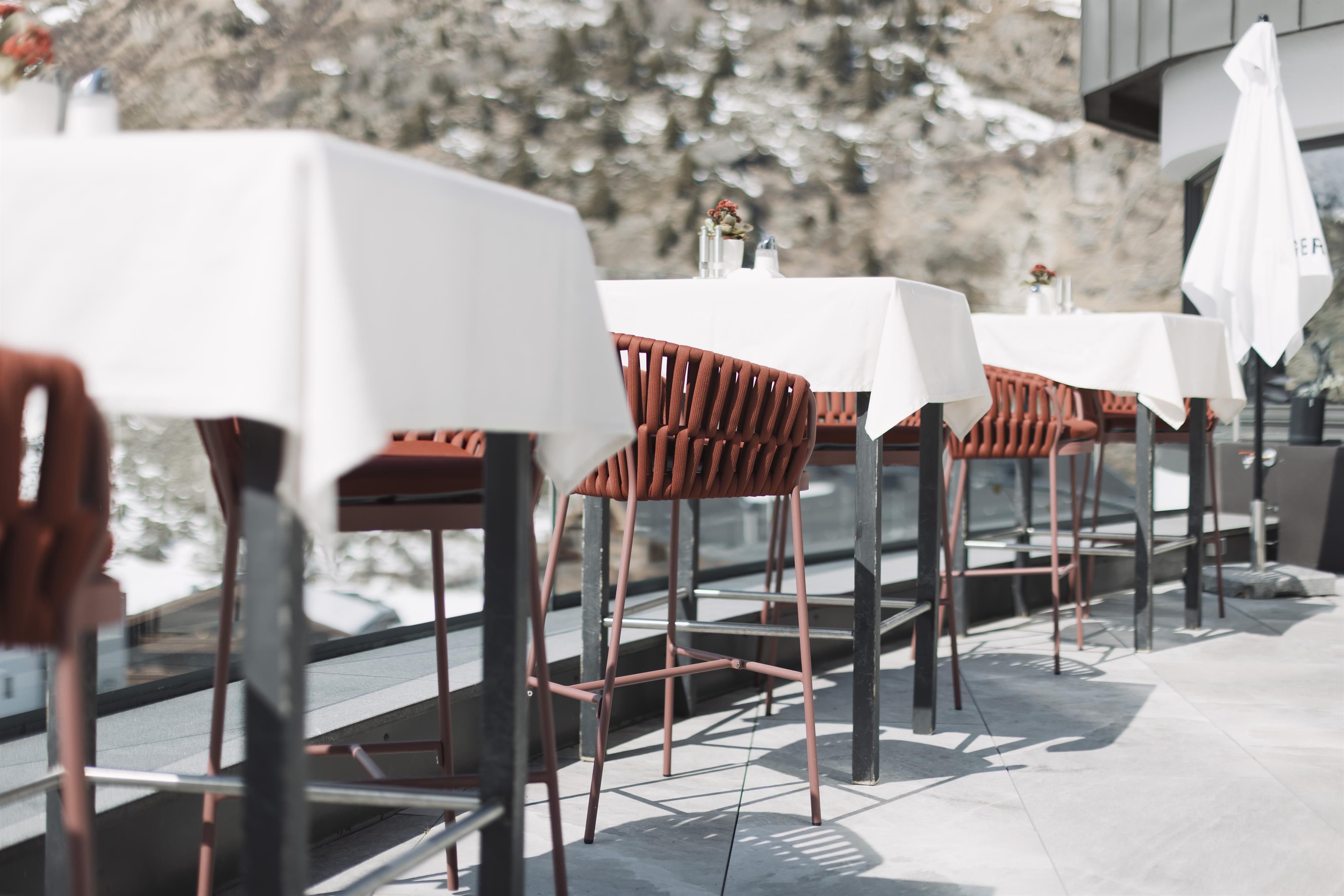 A cozy restaurant terrace with modern tables and chairs. Mountains can be seen in the background.