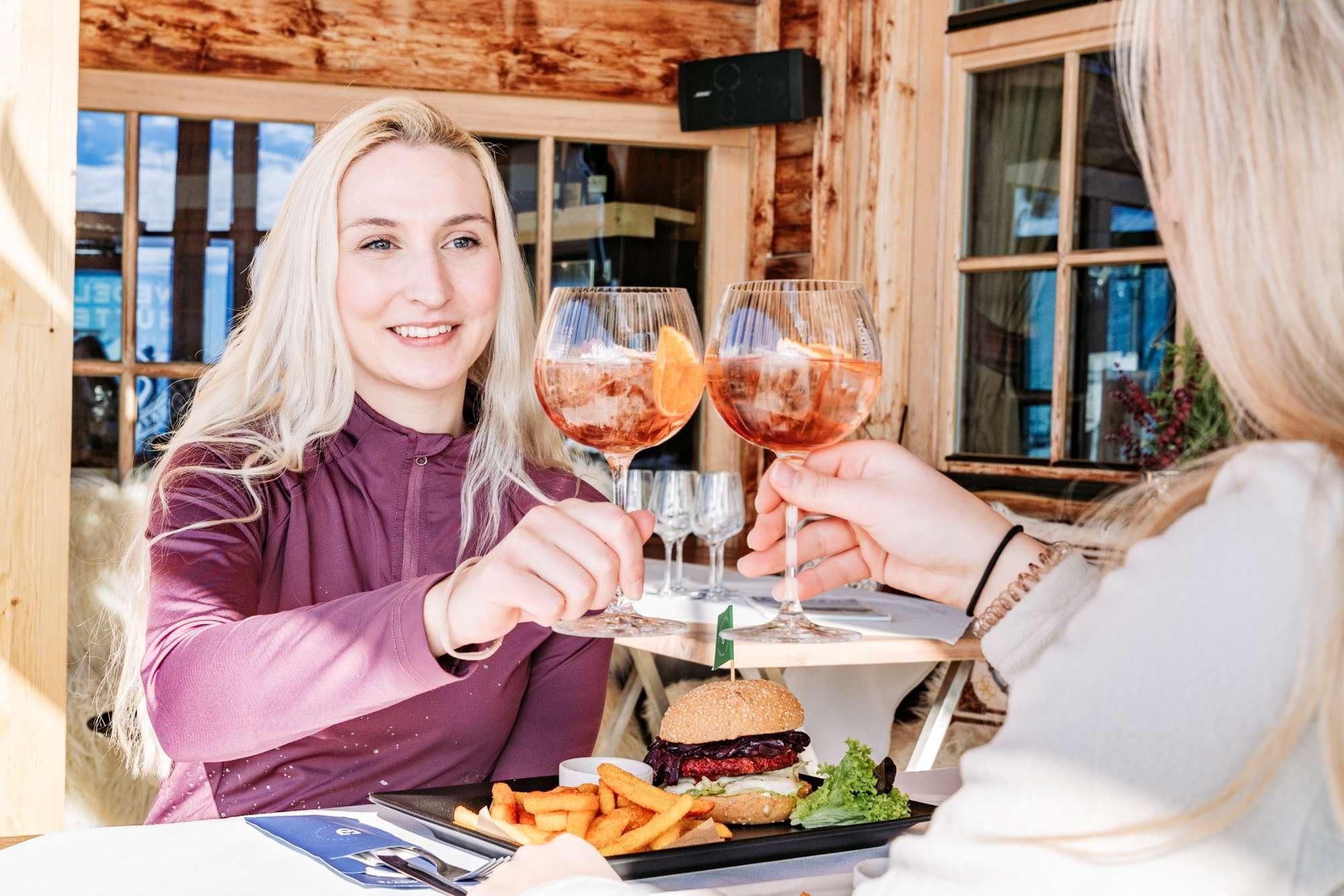 Two women clink glasses and enjoy their meal in a restaurant. On the table lies a burger with fries and fresh vegetables.