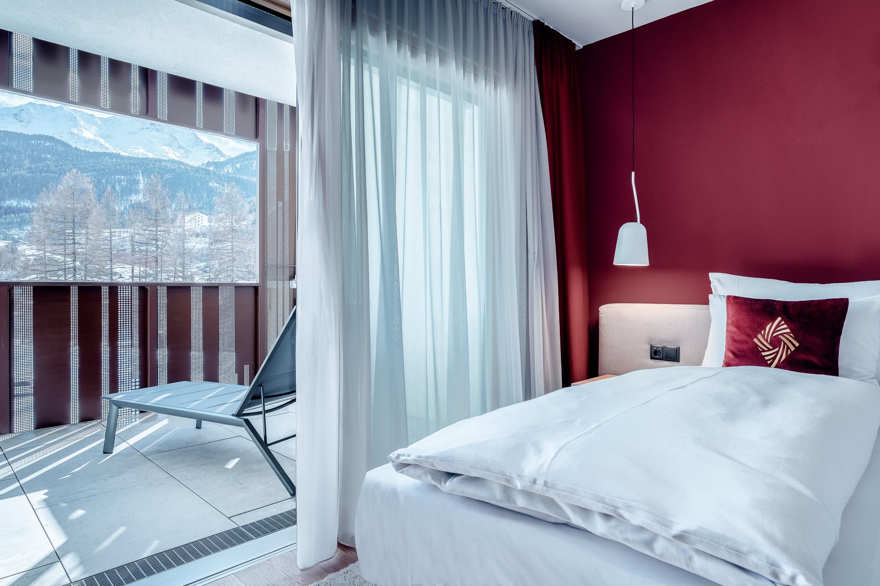 A modern bedroom with red walls and a large window. The room offers a cozy view of the mountains and a private terrace.
