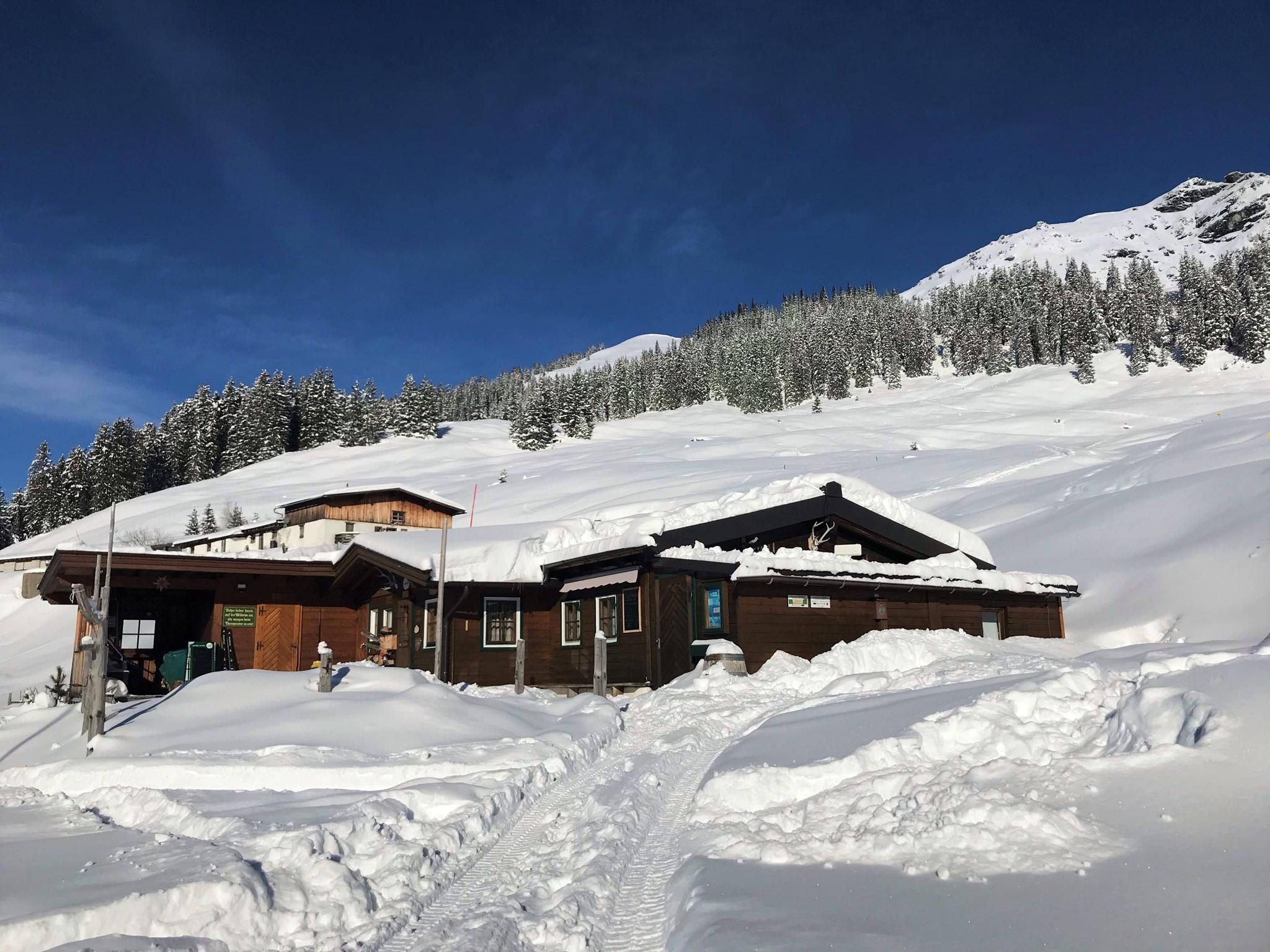 A cozy cabin in the snow, surrounded by snow-covered mountains. The clear blue sky complements the winter landscape.