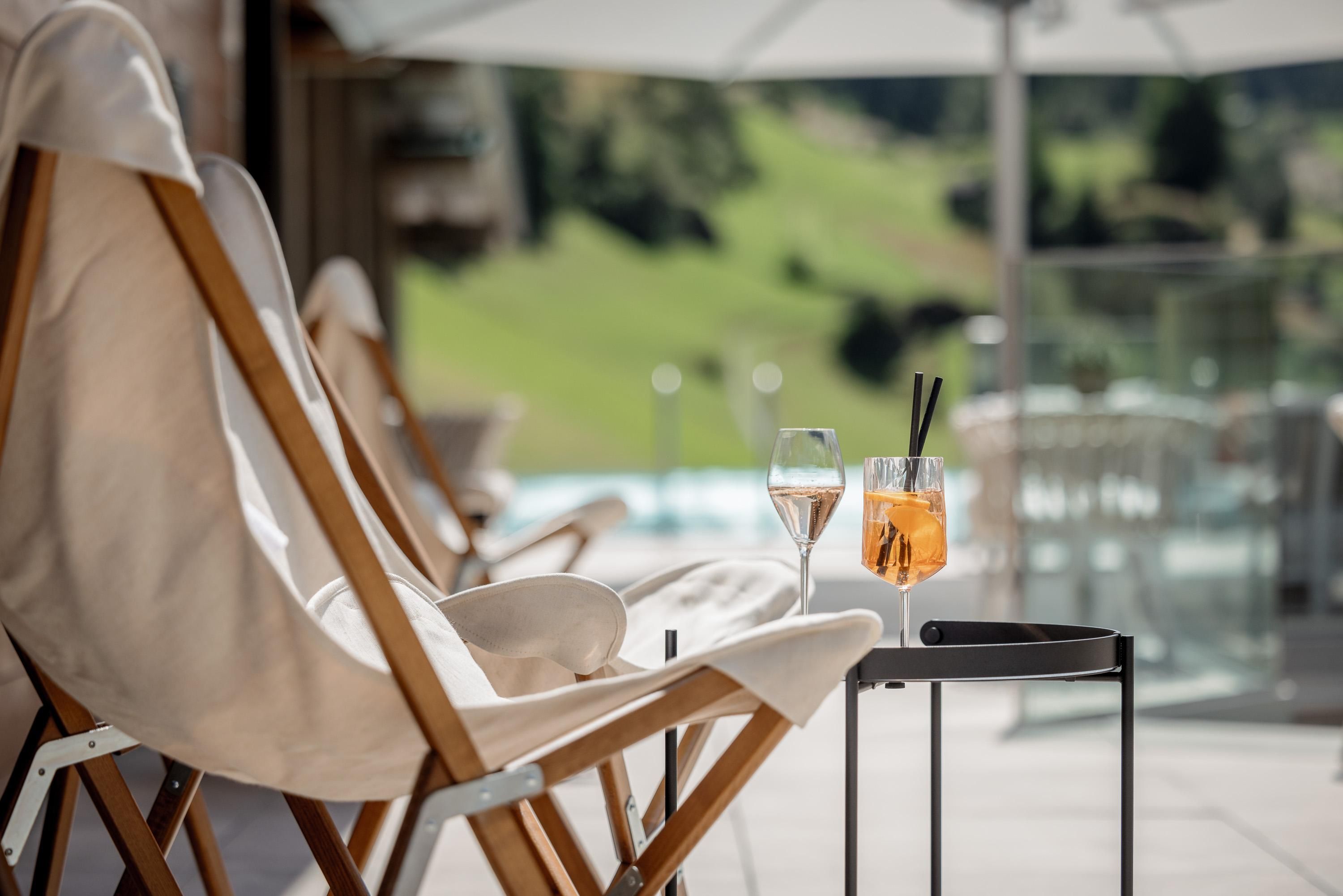 A relaxed area with deck chairs and two glasses of drinks on a table. In the background, a green landscape and a swimming pool are visible.