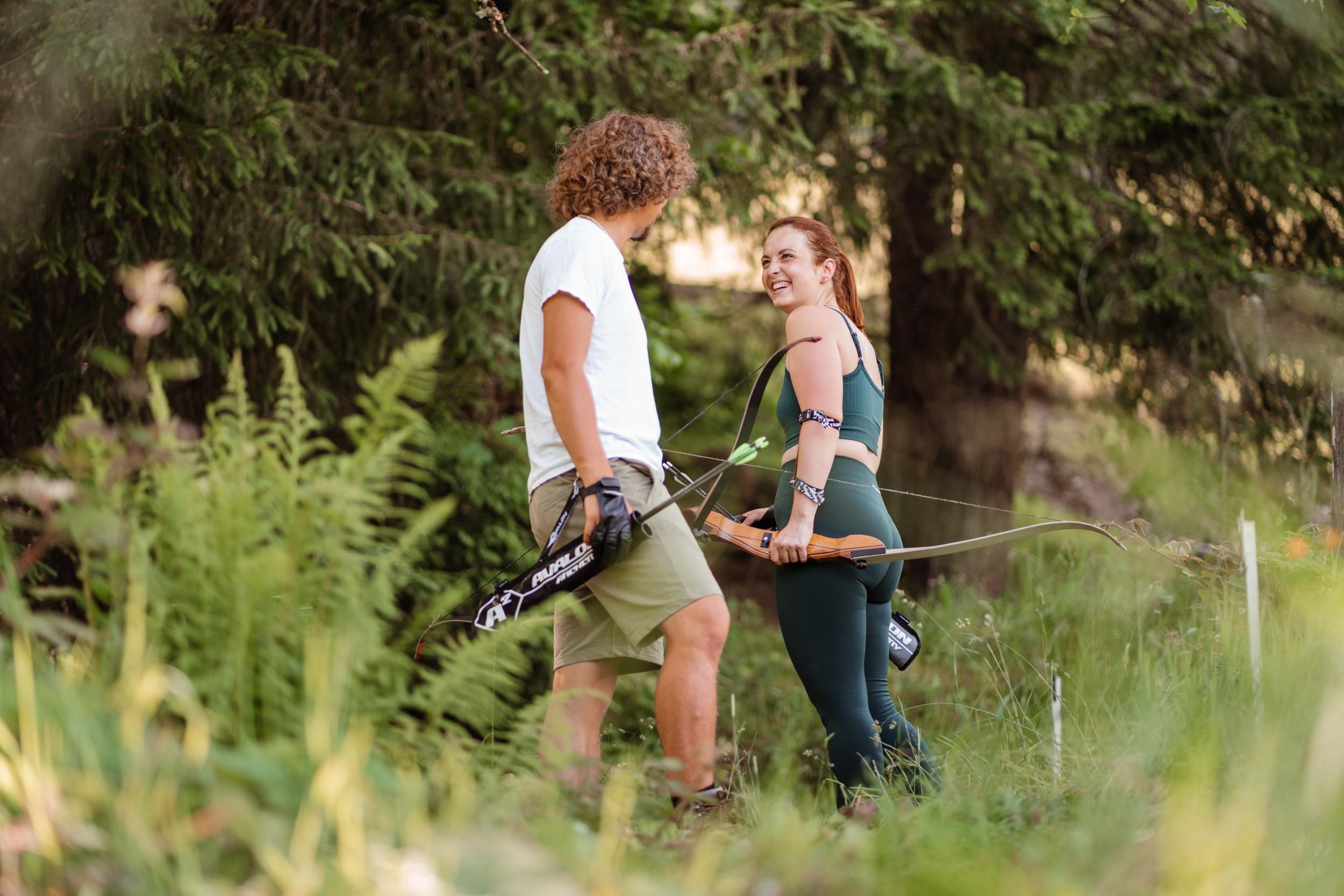 A young couple stands in nature and smiles at each other. They hold bows in their hands and are ready for archery practice.
