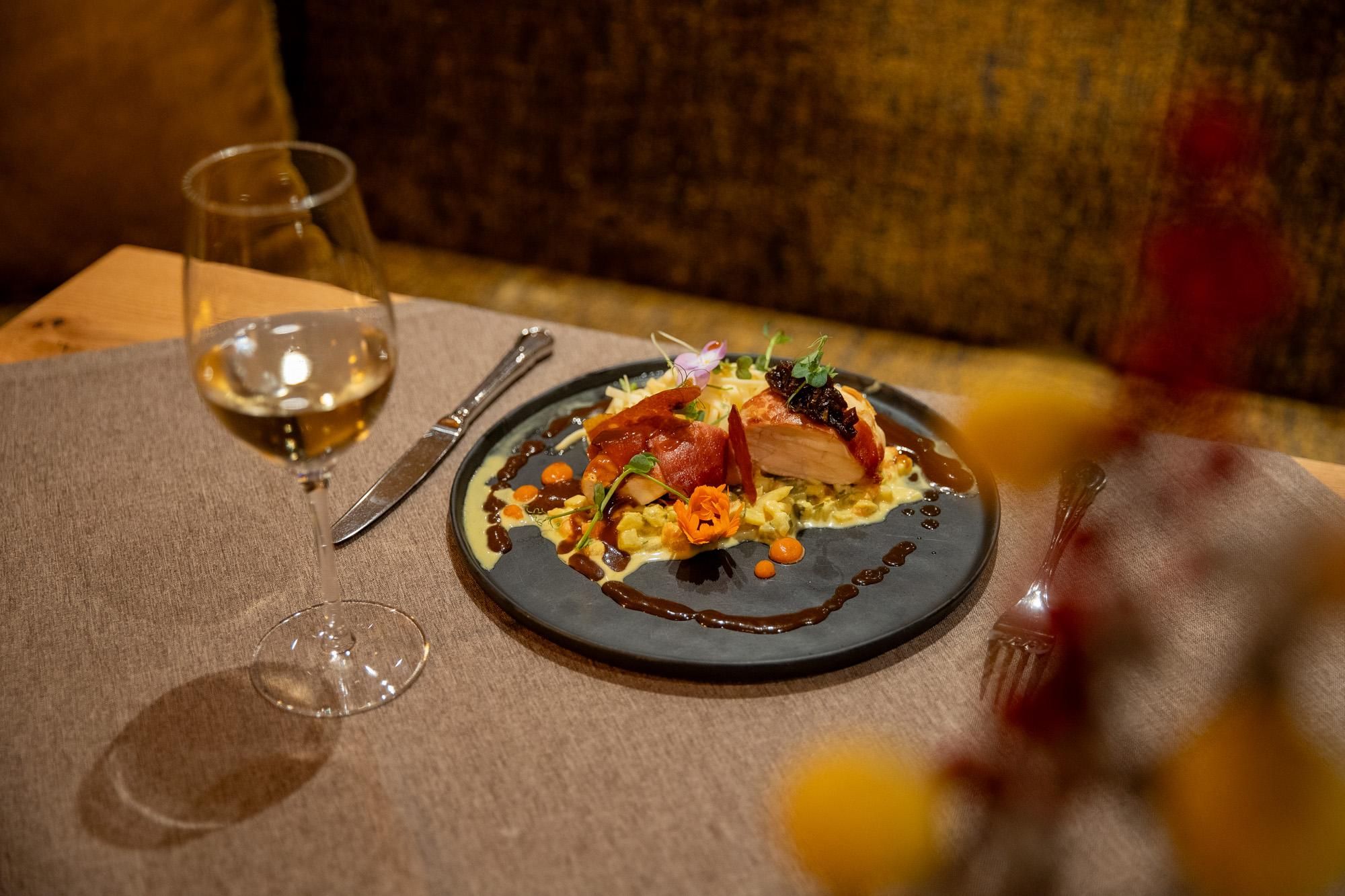An elegant dish on a black plate, garnished with colorful vegetables. Next to it, a glass of white wine is standing on a table with a neutral tablecloth.