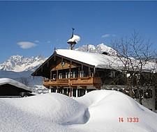 A traditional wooden house in a snowy landscape. In the background, mountains can be seen under a clear blue sky.