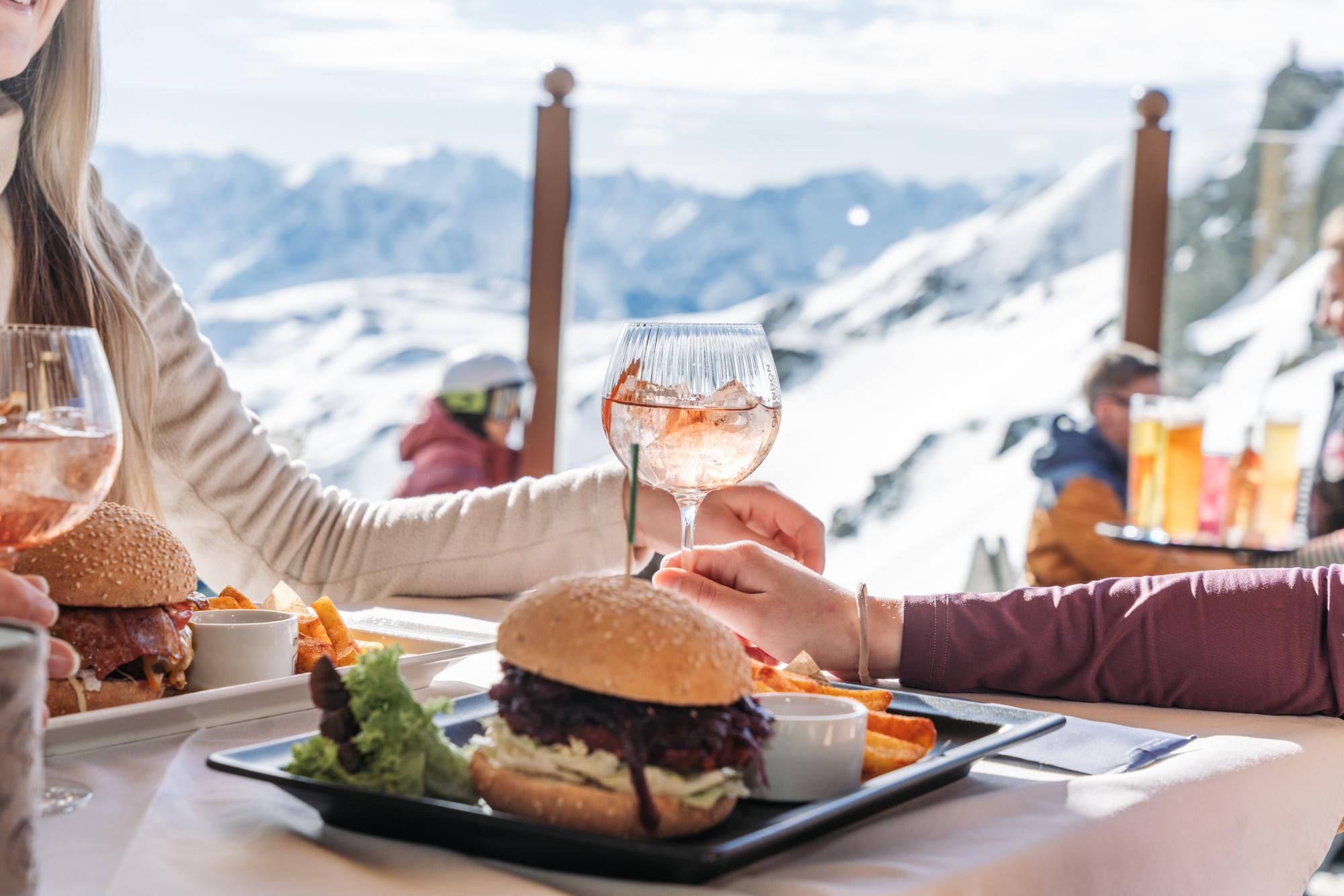 A delicious burger with fries and a glass of wine in a restaurant in the mountains. In the background, you can see snow-covered mountains and a clear sky.