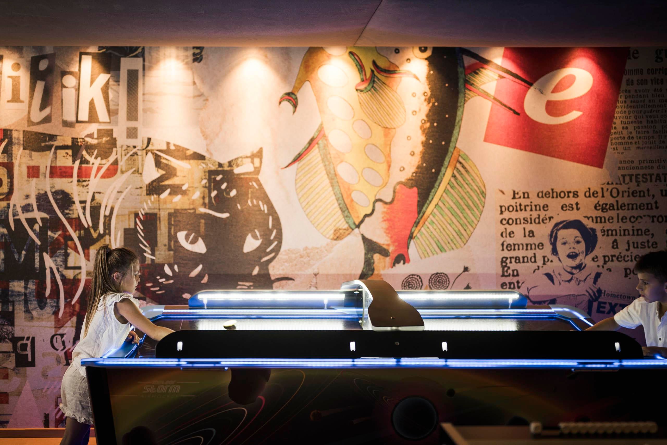 Two children are playing at a modern air hockey table. In the background, a colorful wall with various graphics and texts can be seen.