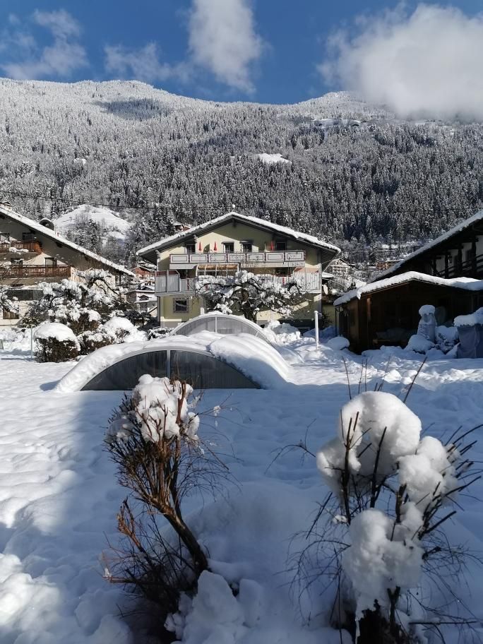 A wintry landscape with snow-covered houses and mountains in the background. The sky is clear and blue, while the snow blankets the surroundings.