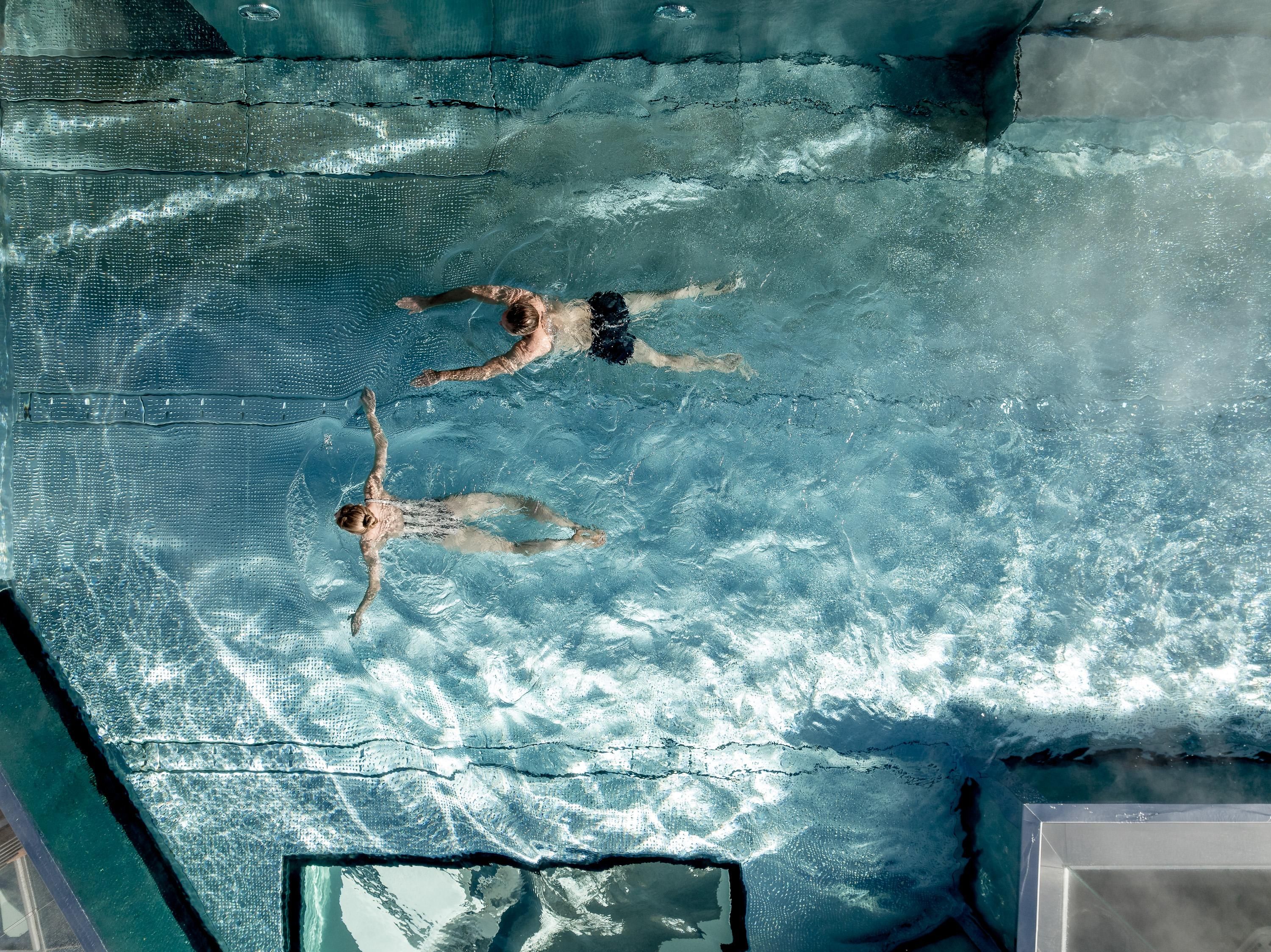 Two people are swimming relaxed in a clear pool. The water surface reflects the light and creates a calm atmosphere.