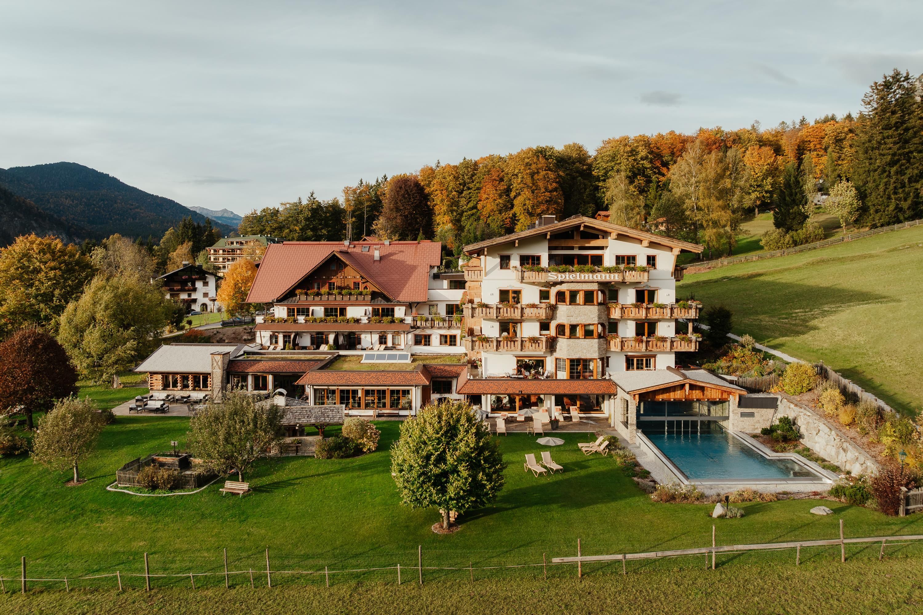 A beautiful hotel in a picturesque landscape with colorful trees in the background. There is a pool and well-maintained gardens.