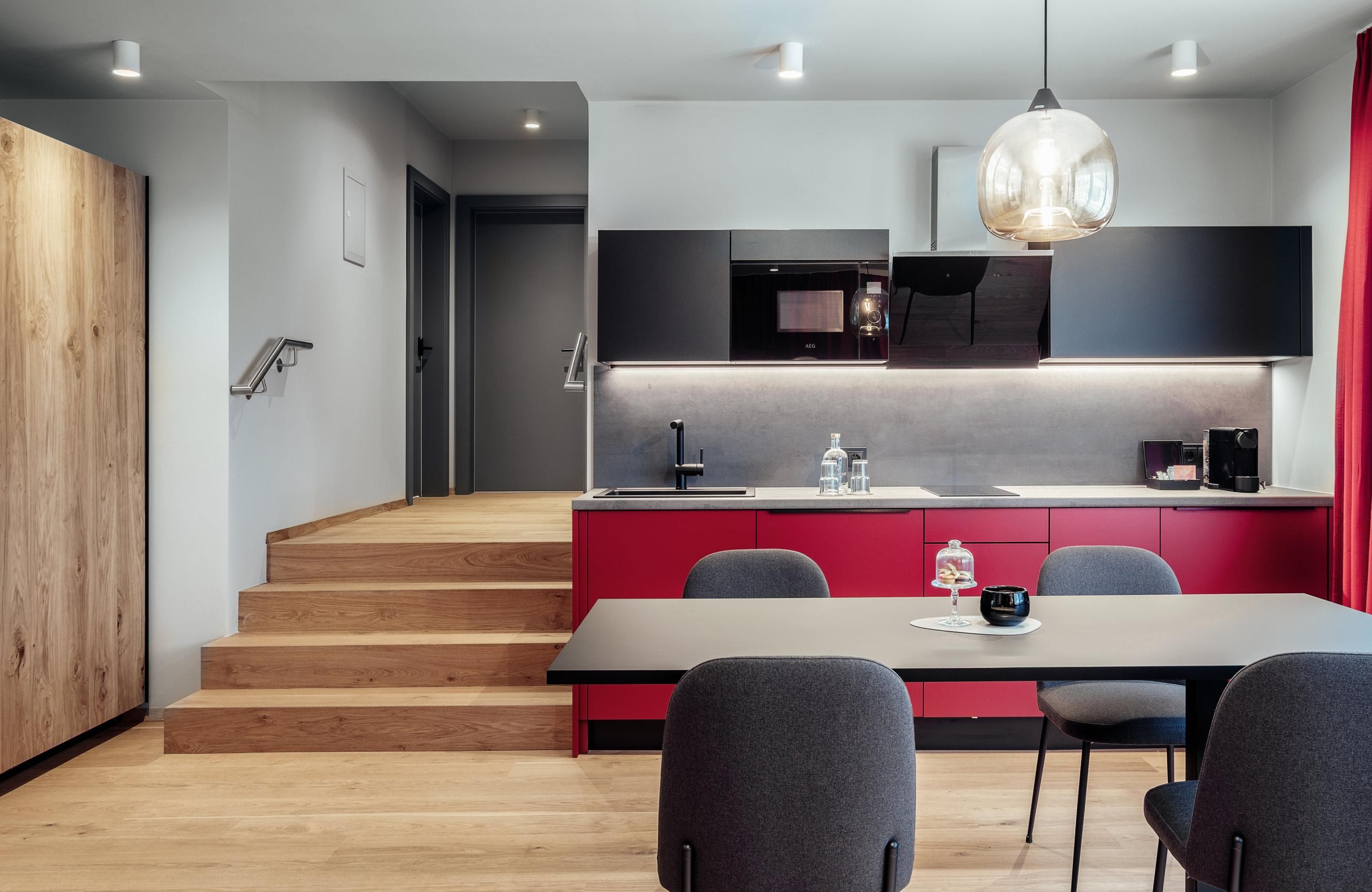 A modern kitchen with red cabinets and a large dining table. The room has a bright wooden floor and a minimalist decor.