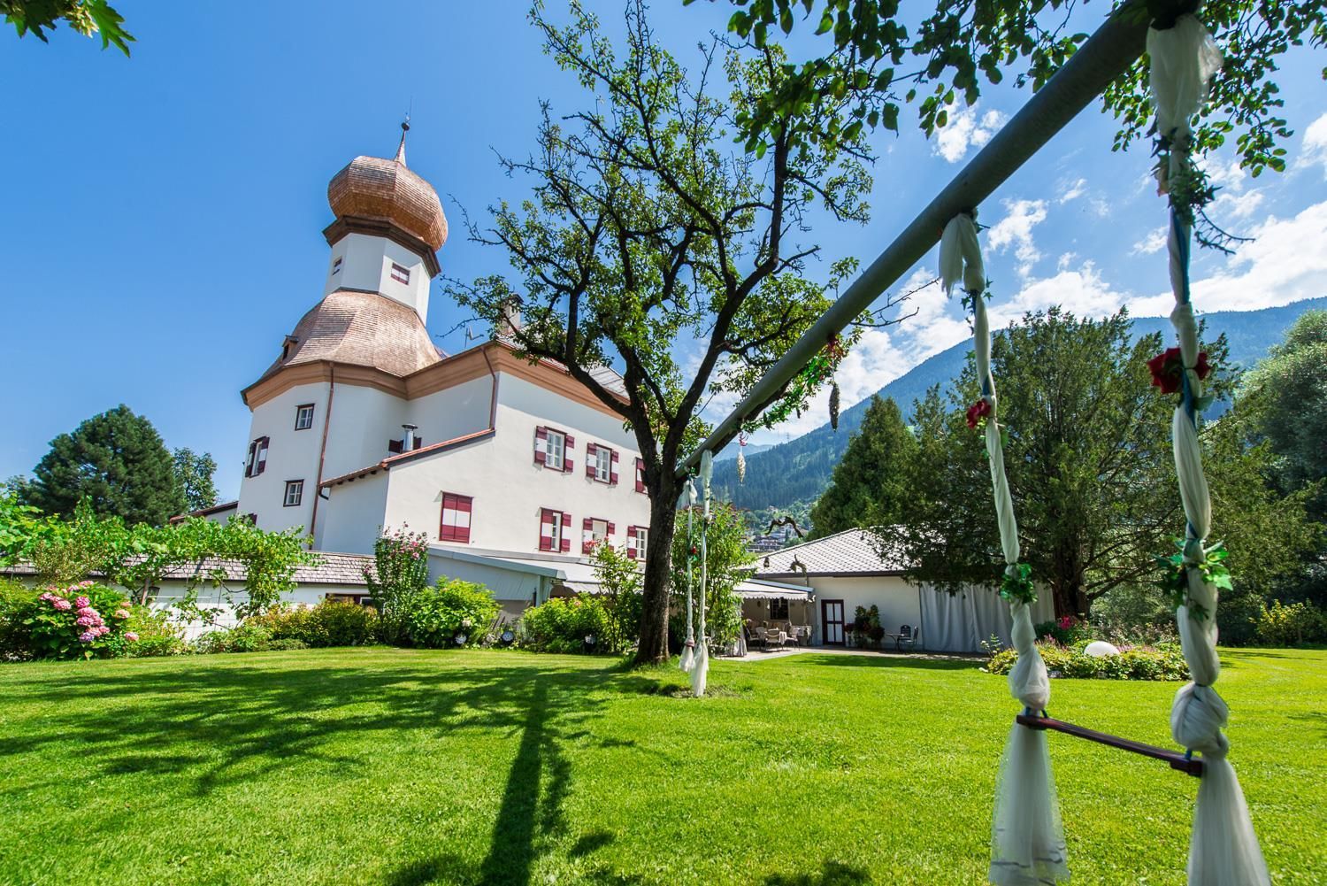 A beautiful building with a distinctive dome is located in a well-maintained garden. In the foreground, a swing can be seen under a tree.