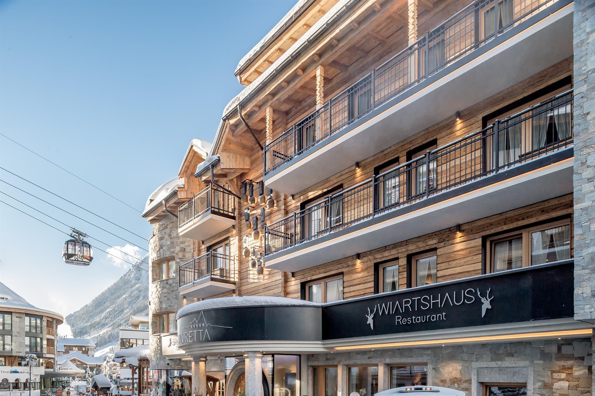 A modern building with a wooden and stone facade in a mountain landscape. In the foreground, the restaurant "Wiartshaus" can be seen, and in the background, snow-covered mountains.