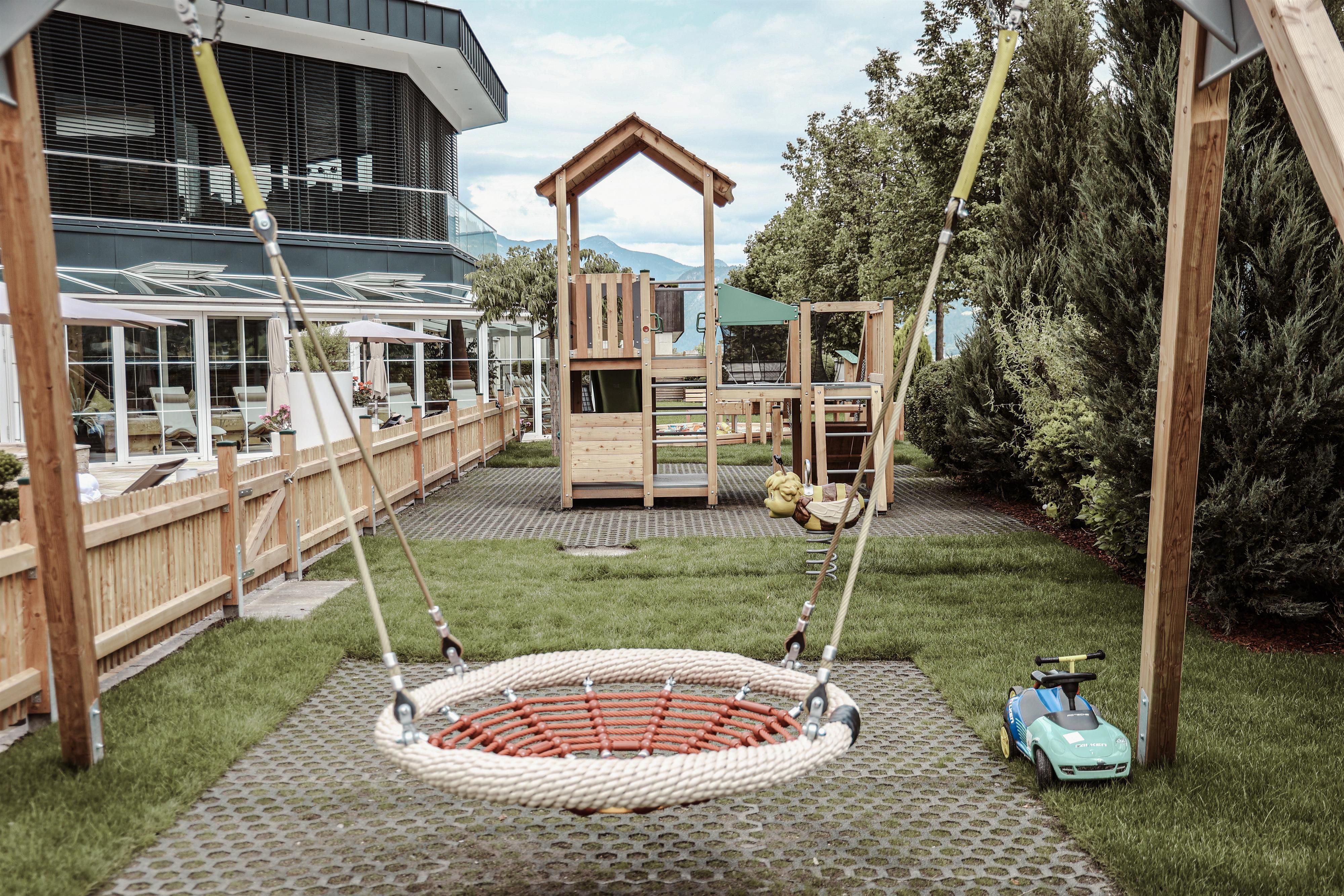 A playground with a large net swing and a play tower structure. In the background, there is green grass and some trees.