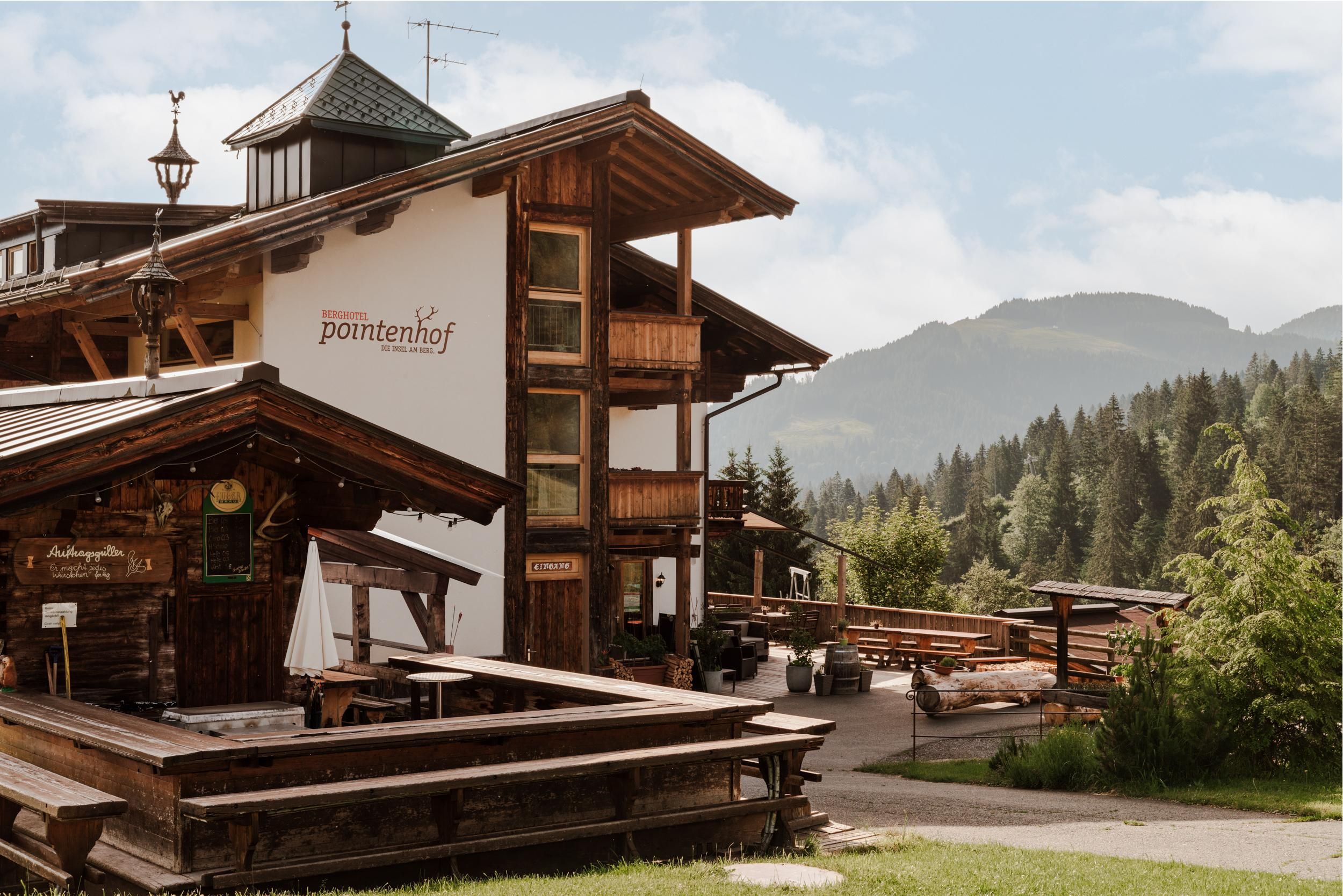 A charming wooden building named "Pontenhof" surrounded by green trees and mountains. The terrace is inviting with outdoor seating.