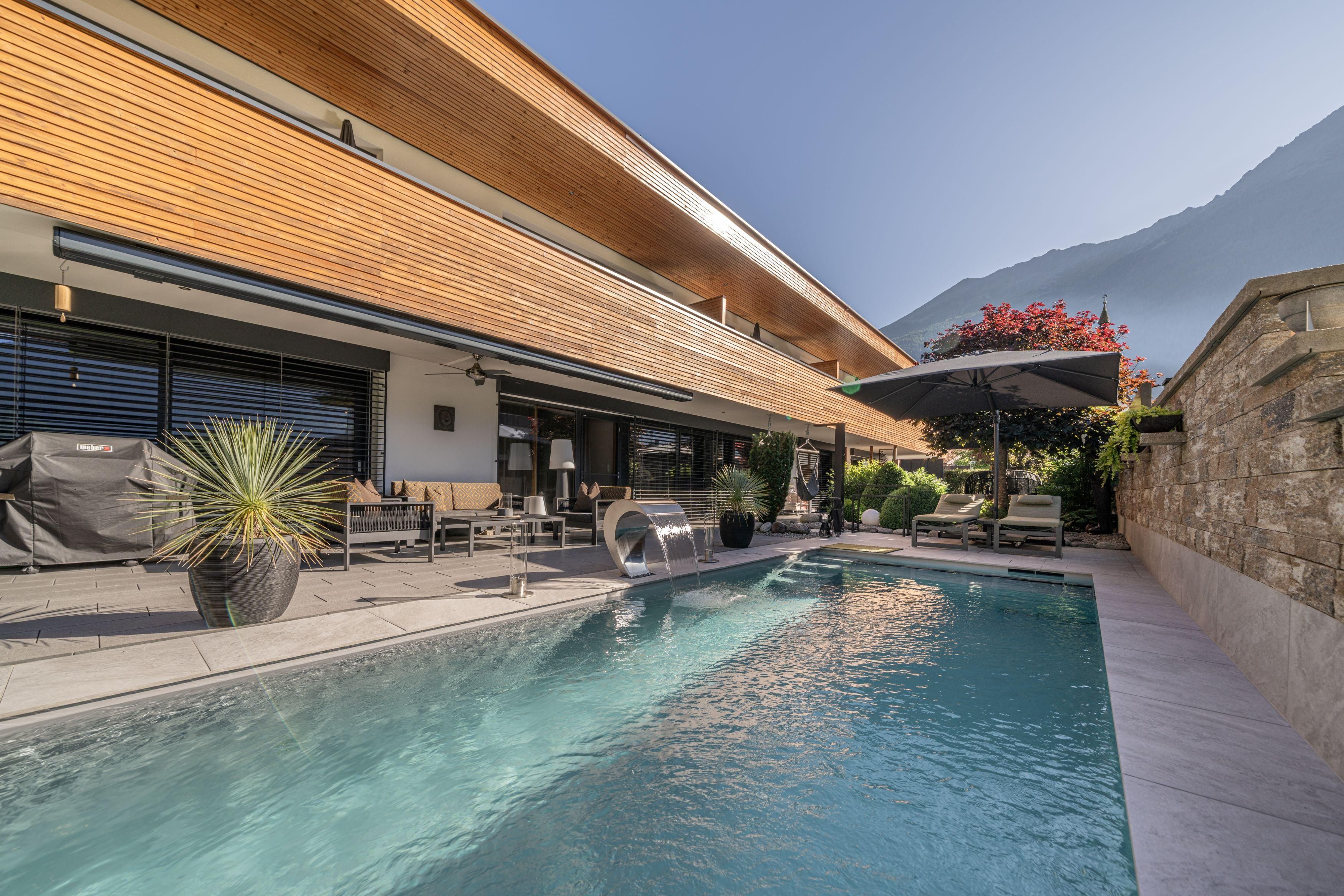 A modern outdoor area with a swimming pool and cozy seating. In the background, green plants and a mountain landscape can be seen.