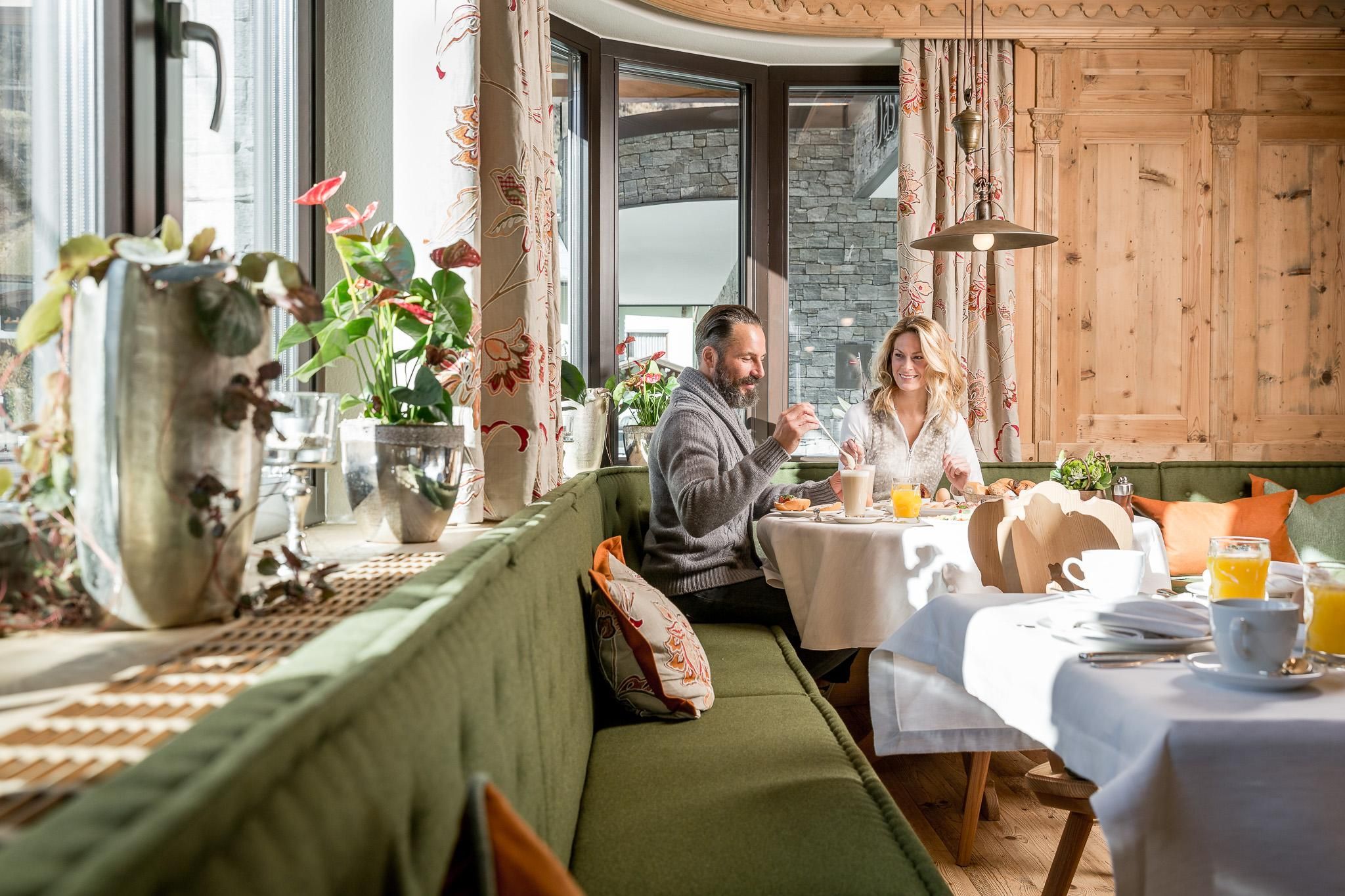 A cozy breakfast room with a green sofa and a table set with food and drinks. Two people are sitting comfortably and enjoying their meal.