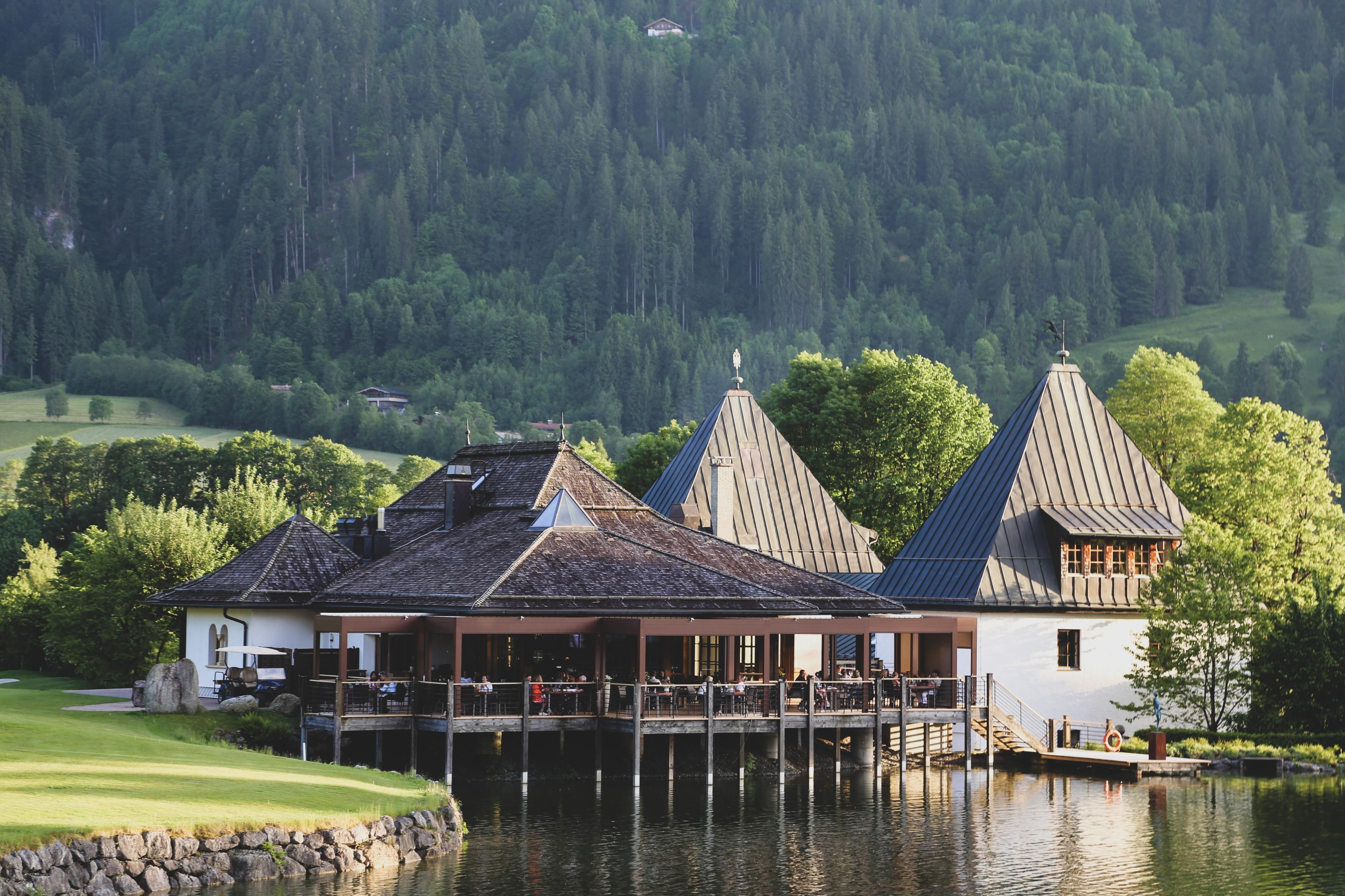 A picturesque building by the water, surrounded by green trees and mountains. The architecture features striking, pointed roofs and an inviting terrace.
