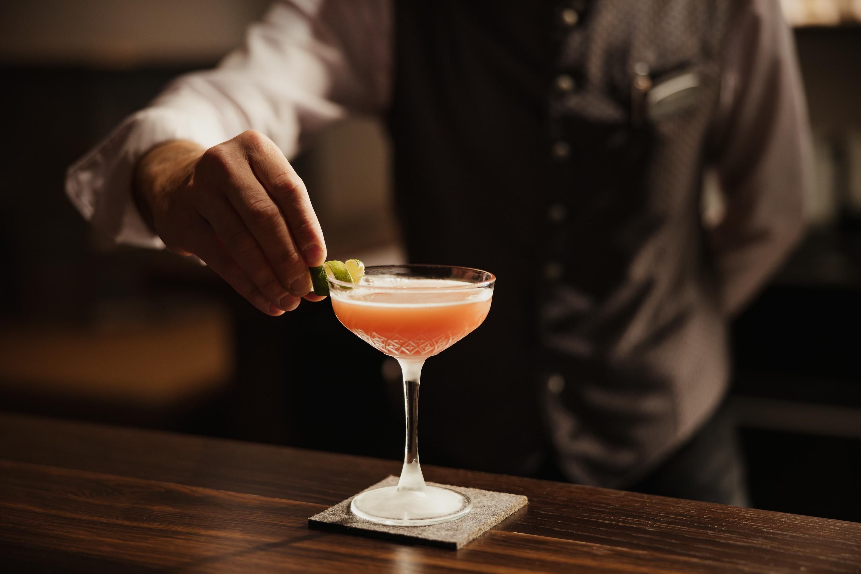 A bartender garnishes a cocktail in an elegant glass. The drink has a pink color and is decorated with a slice of lime.