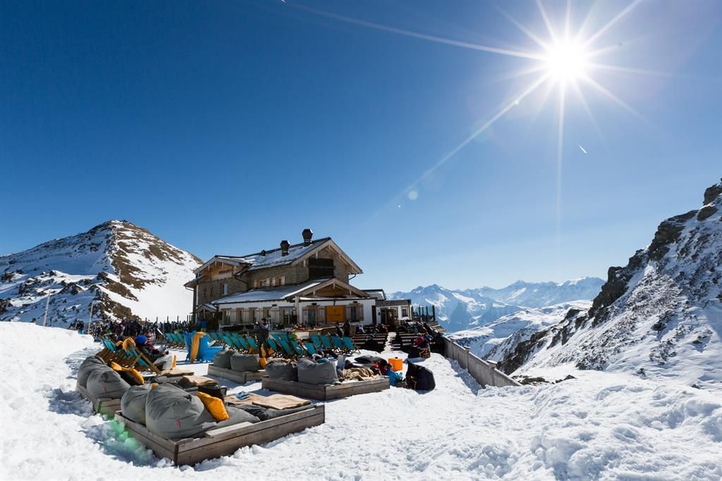 A picturesque mountain house in snow-covered mountains under a clear blue sky. Cozy seating is located outdoors, perfect for relaxing in the sun.