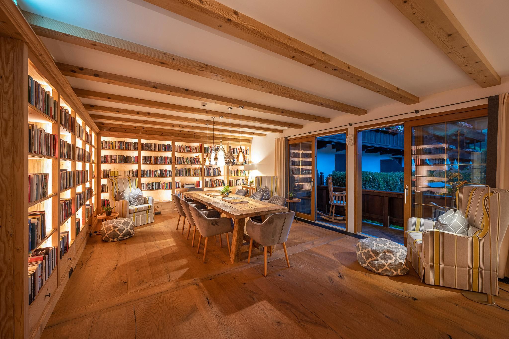 A cozy room with wooden shelves full of books and a large table in the middle. Large windows let in light and offer a view outside.